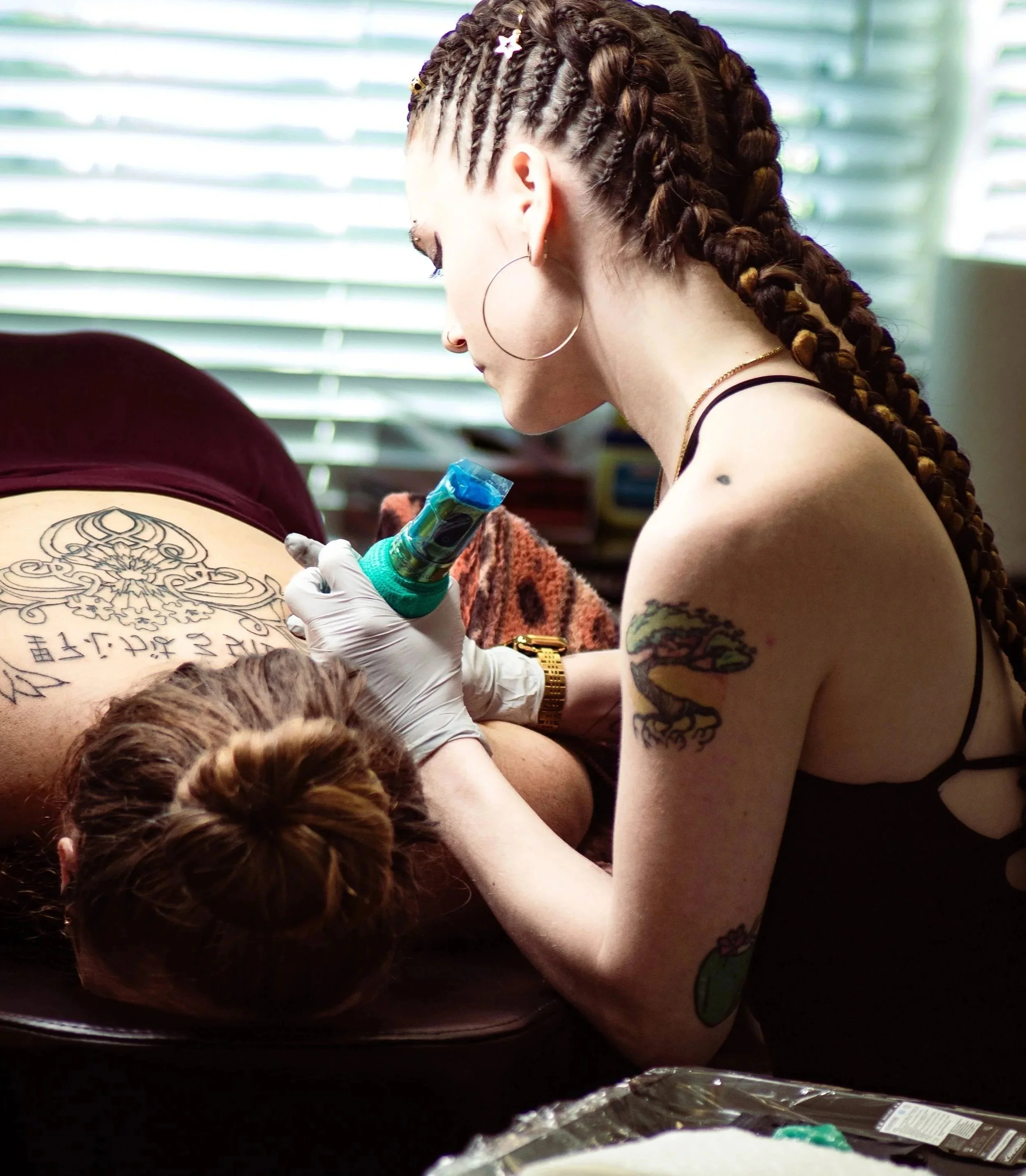 A woman with tattoos on her arms and large hoop earrings, with braided hair decorated with beads, is tattooing a large, intricate black tattoo on someone's back. She is wearing white gloves and holding a tattoo machine. The setting appears to be a bright room with green and white blinds in the background.