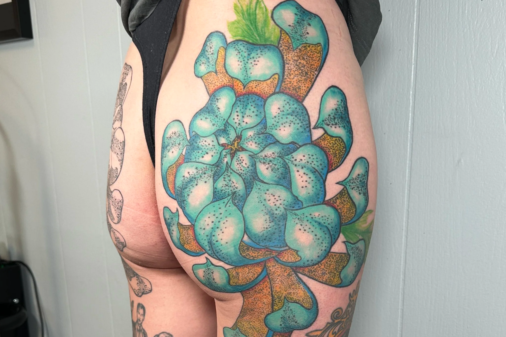 A colorful tattoo of a large peony flower on a person's upper arm and shoulder, featuring blue, green, orange, and yellow hues, along with black outlines.