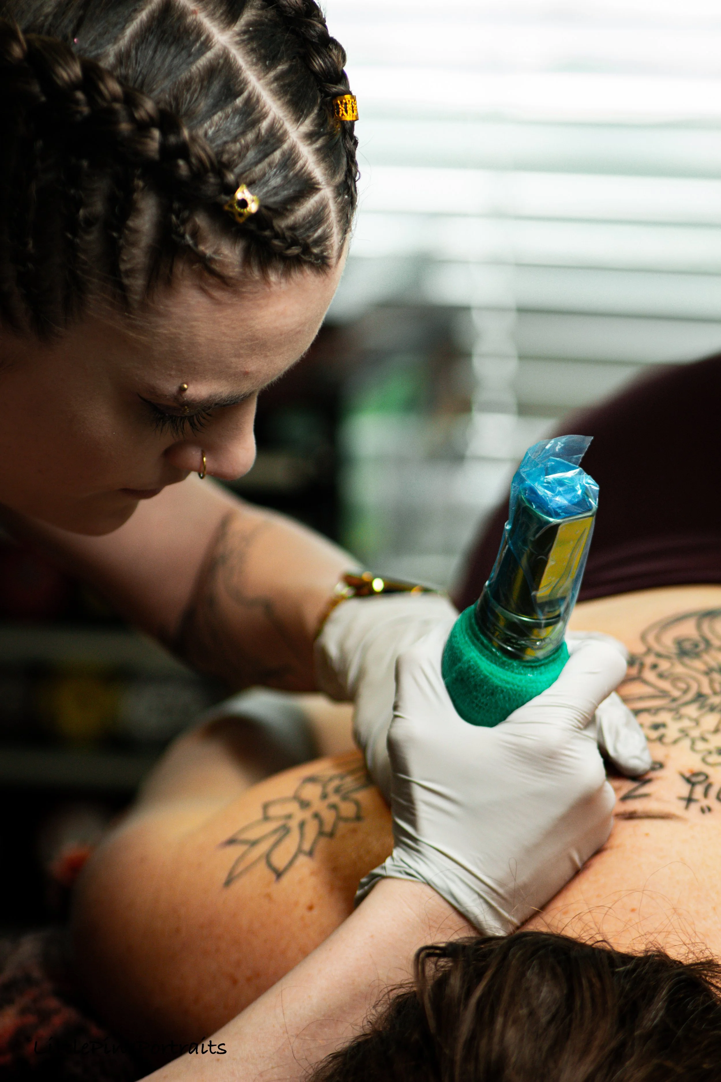A person with braided hair and facial piercings is getting a tattoo on their arm by an artist wearing gloves, in a tattoo studio.