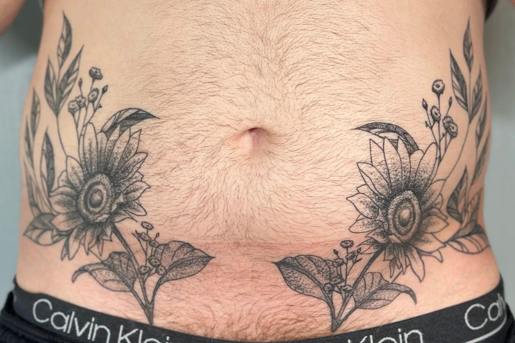Tattoo of sunflowers and leaves around a navel on a person's abdomen.
