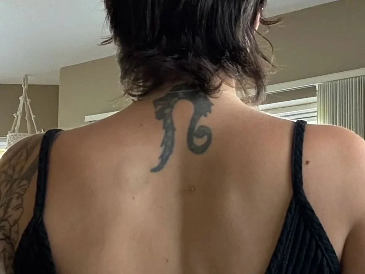 Back of a woman with shoulder-length dark hair, black tank top, and a tattoo on her upper back near the neck.
