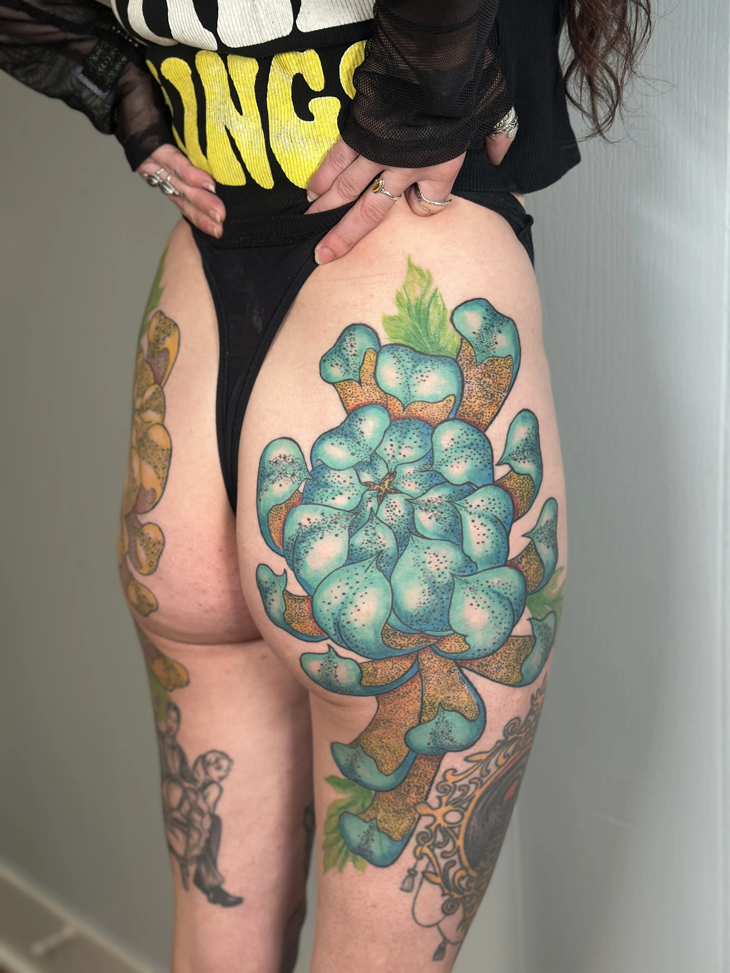 A person with multiple tattoos, including a large blue-green succulent plant on the thigh, wearing black underwear and a black shirt with yellow text, has their hand resting on their hip, displaying rings and jewelry.