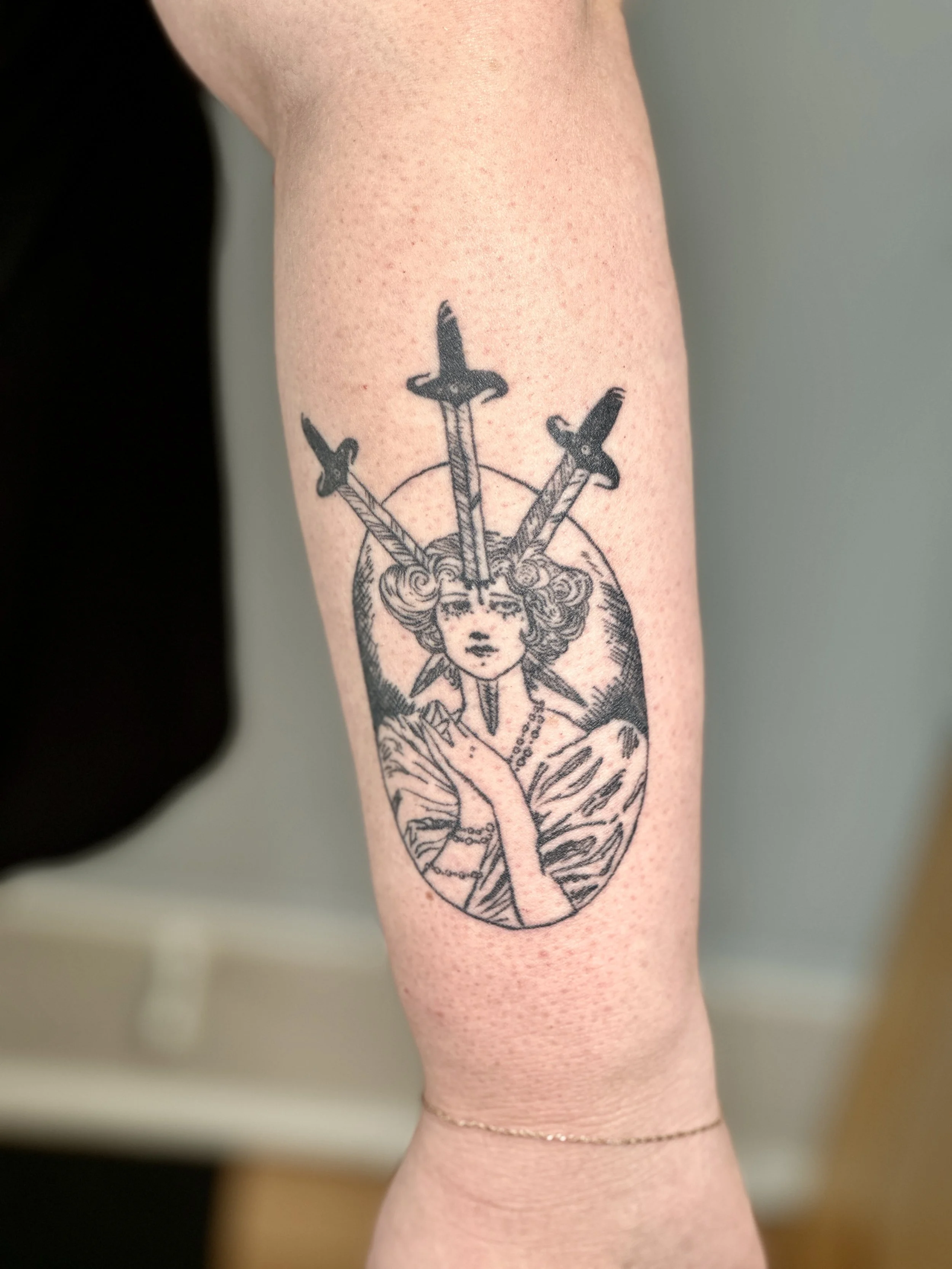 Tattoo of a woman with three swords piercing her head, surrounded by a circular design, on a person's arm.