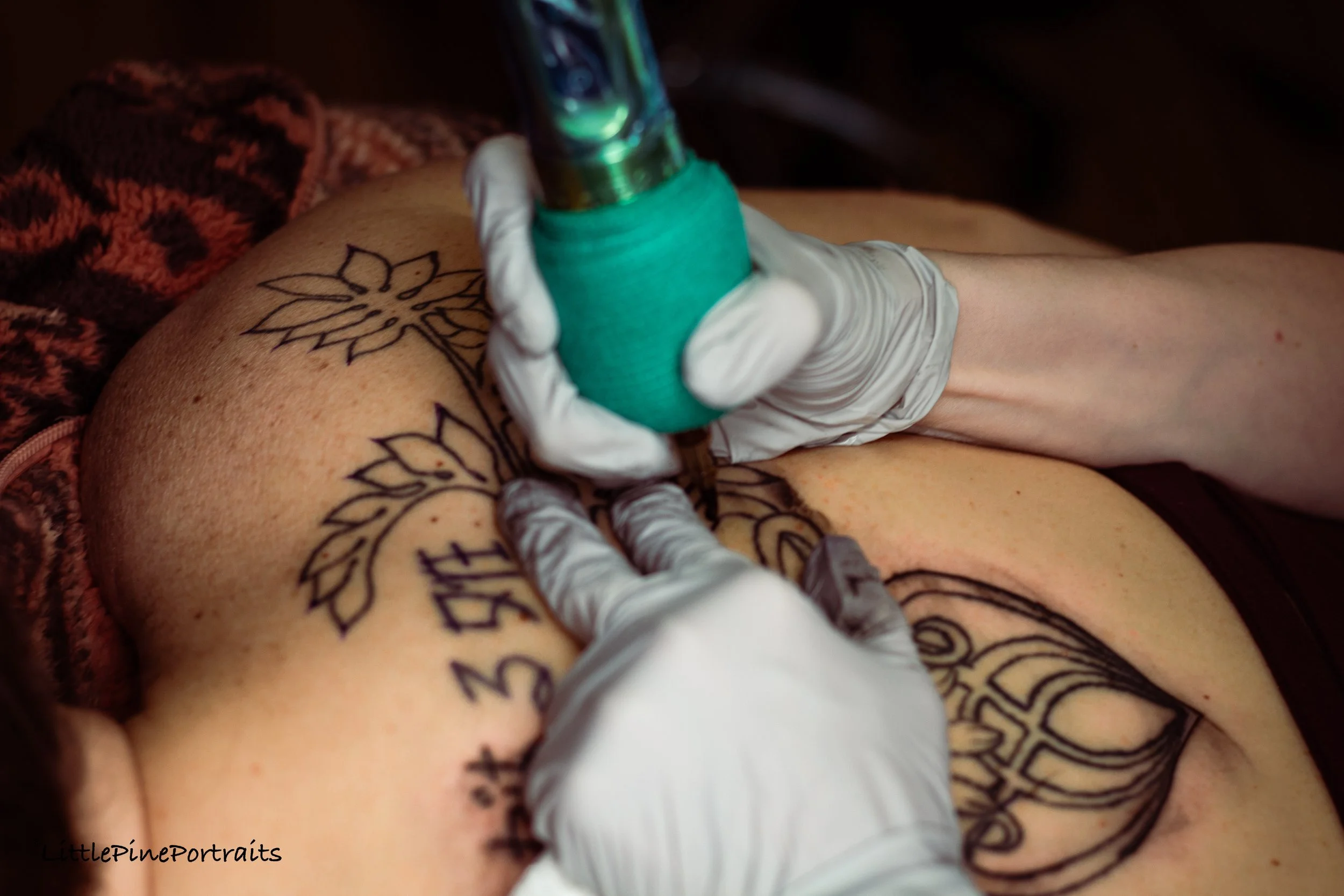 A tattoo artist is creating a floral tattoo with black ink on a person's skin using a tattoo machine. The person's skin has multiple black outline tattoos, and the artist is wearing white gloves. A maroon, black, and white patterned fabric is visible