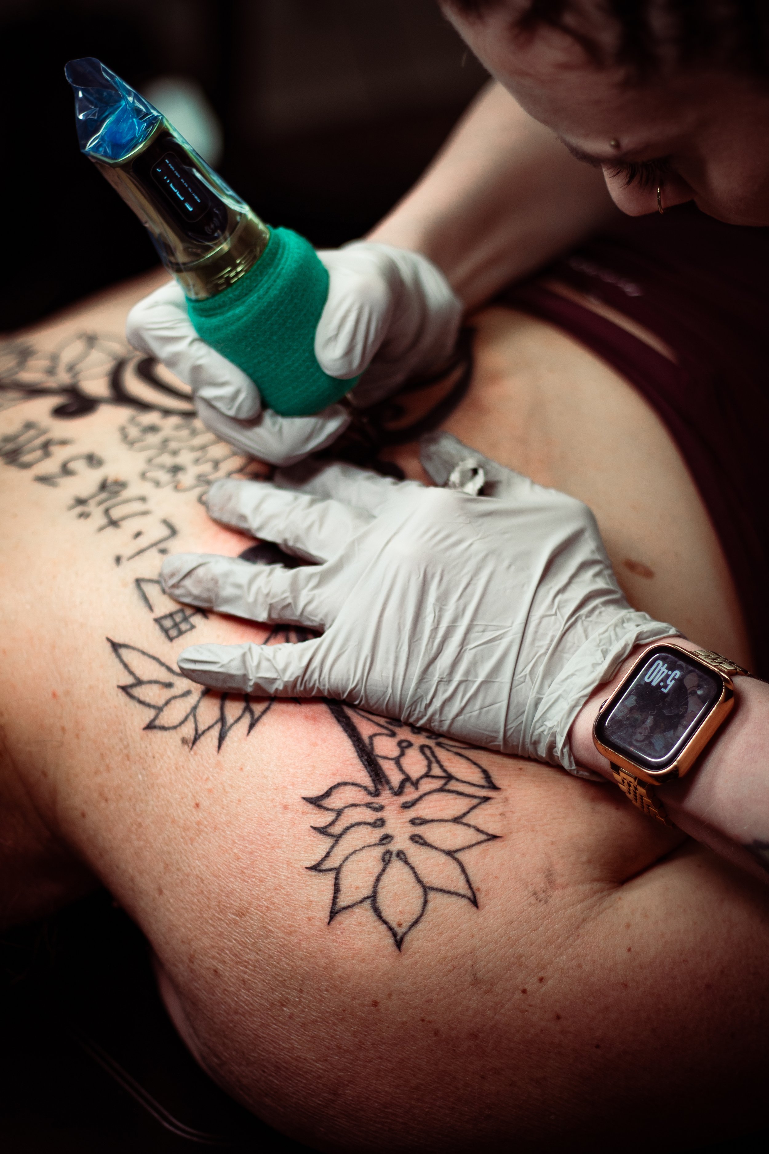 A person with tattoos on their skin is getting a tattoo. They are wearing a smartwatch on their wrist and a smartwatch with a yellow band. The tattoo is of floral designs. The person is using a tattoo machine, which is being held by a hand wearing a 