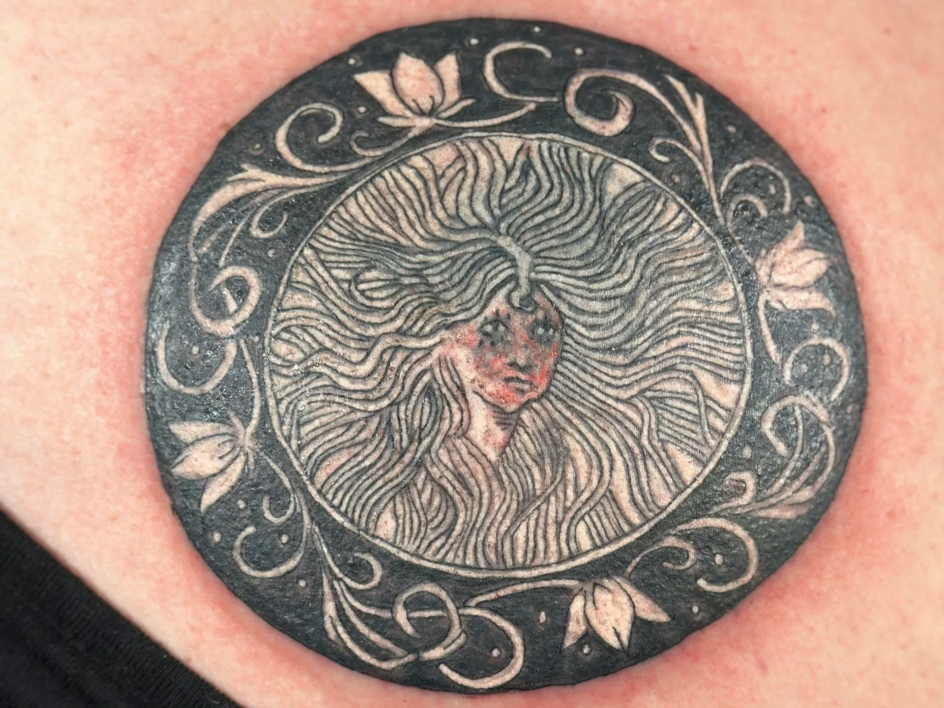Close-up of a black tattoo featuring a stylized face of a woman with flowing hair, surrounded by ornate decorative elements in circular design.