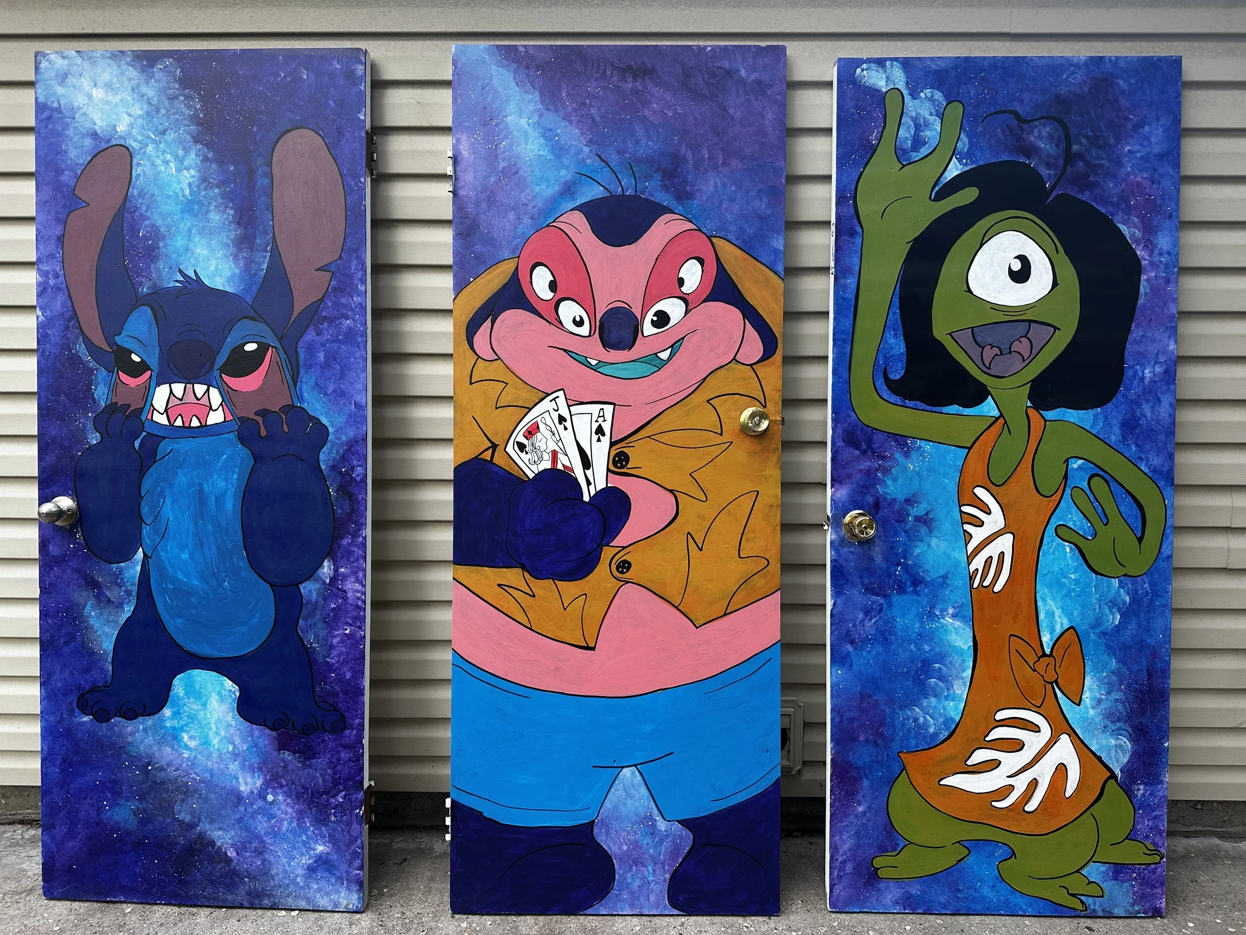 Colorful painted panels featuring cartoon characters on a gray horizontal wall. The characters include a blue creature with long ears, a pink creature with multiple eyes holding playing cards, and a green creature with one eye and large antennae.