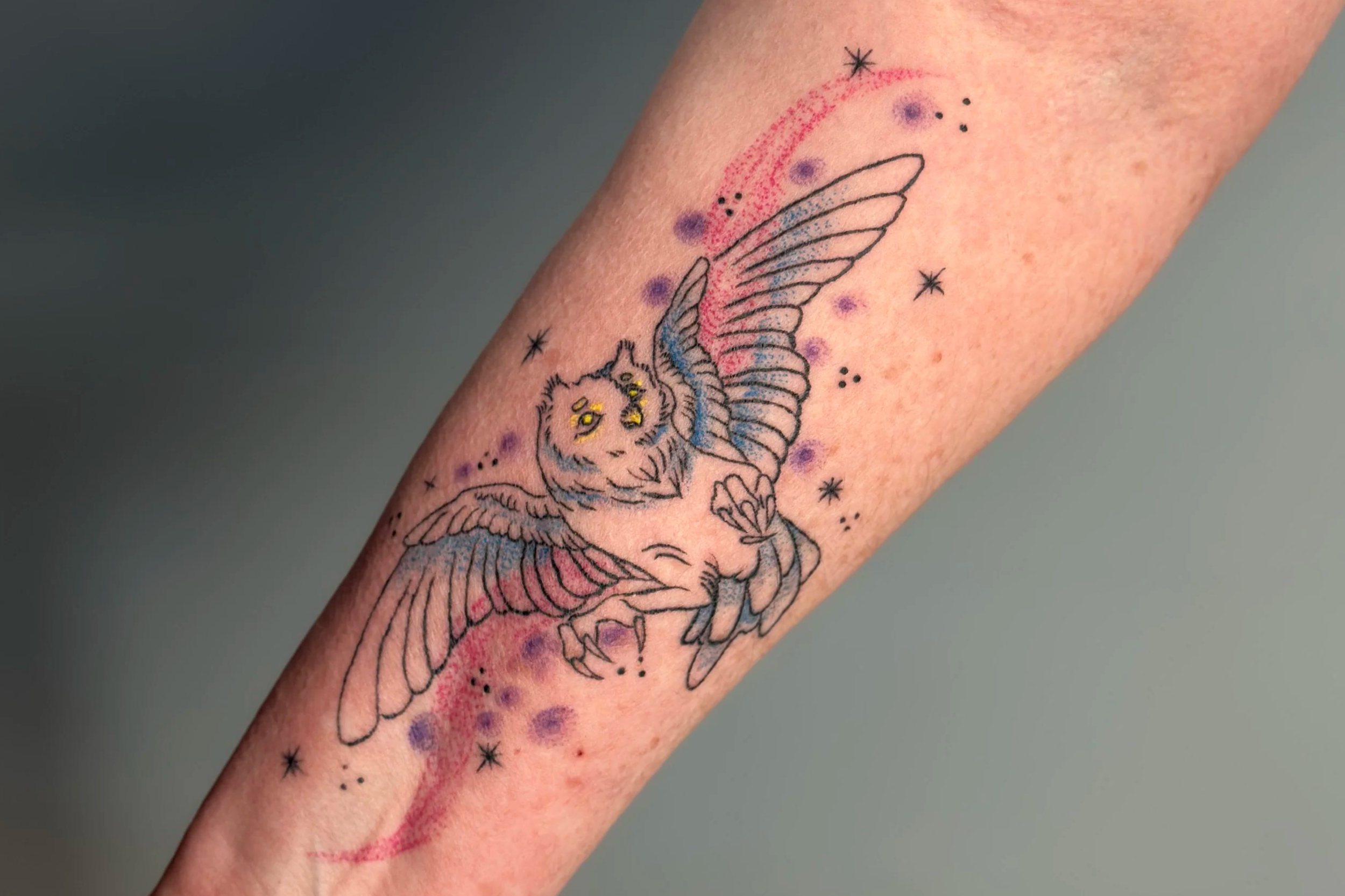 Tattoo of a flying owl with colorful accents, surrounded by small decorative stars and dots on a person's arm.