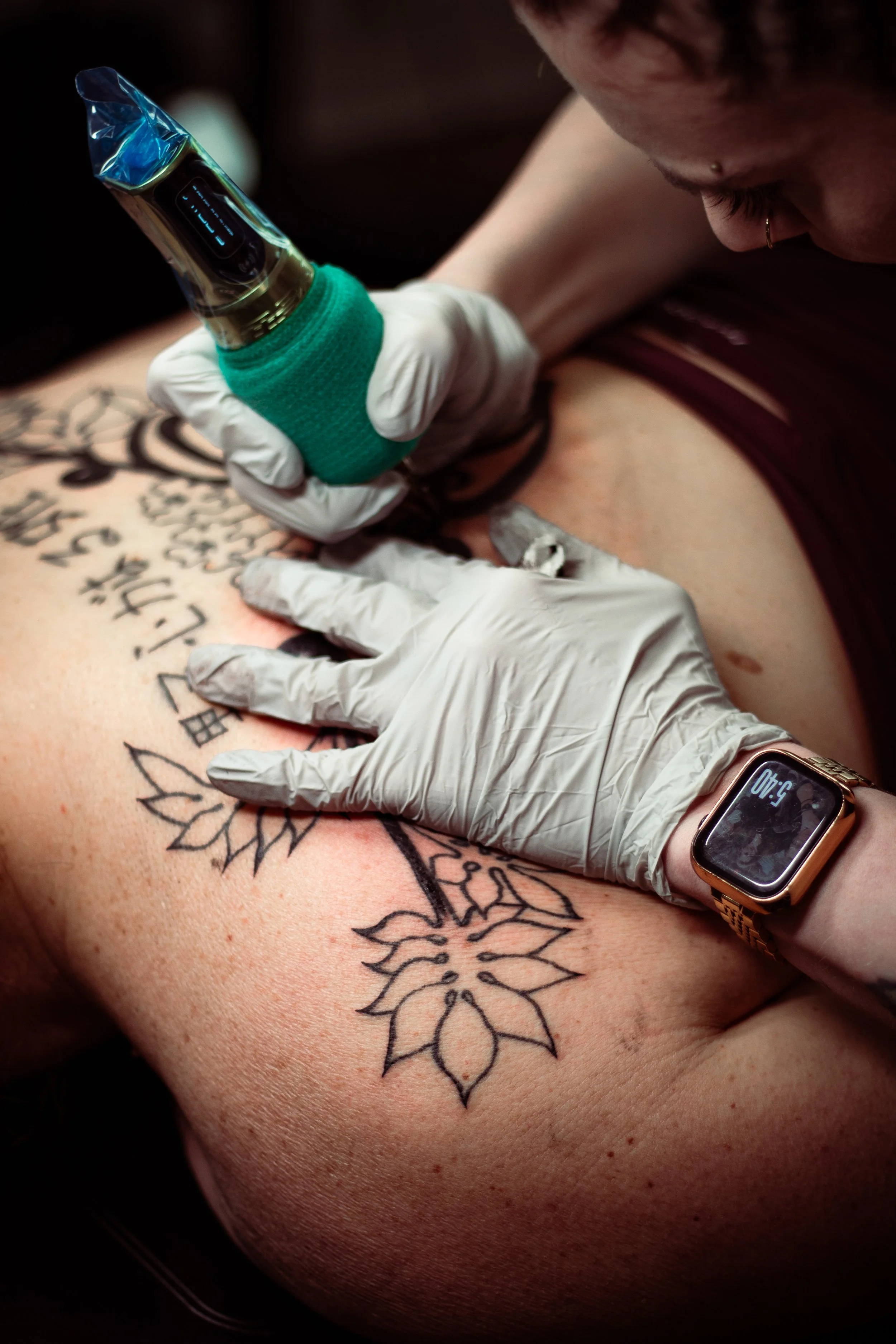 Person with a tattoo of flowers on their back getting a tattoo from an artist wearing gloves, a tattoo machine, and a smartwatch showing 3:40.
