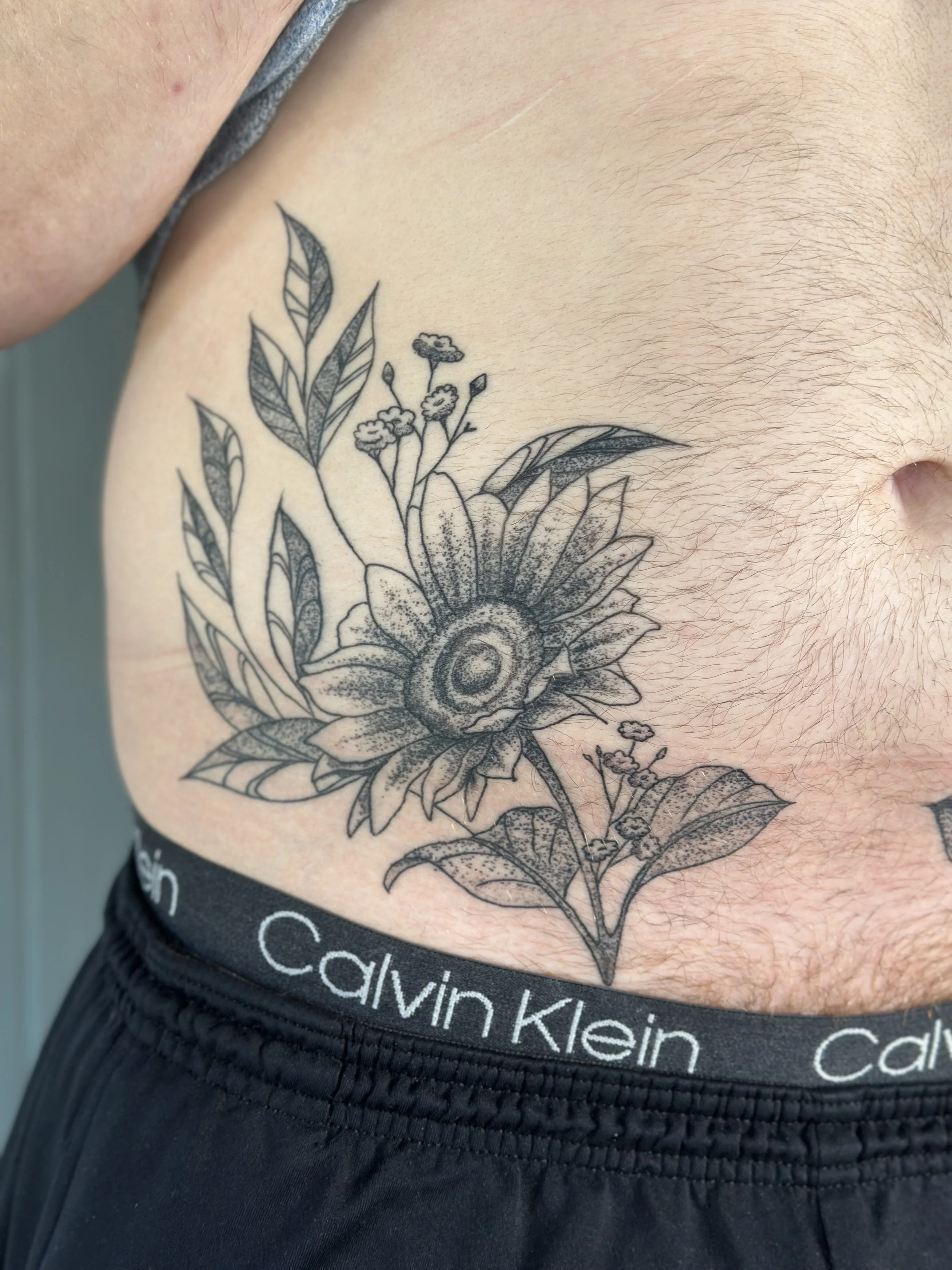Close-up of a person's lower torso showing a floral tattoo on the side of the waist, partially covered by Calvin Klein underwear.