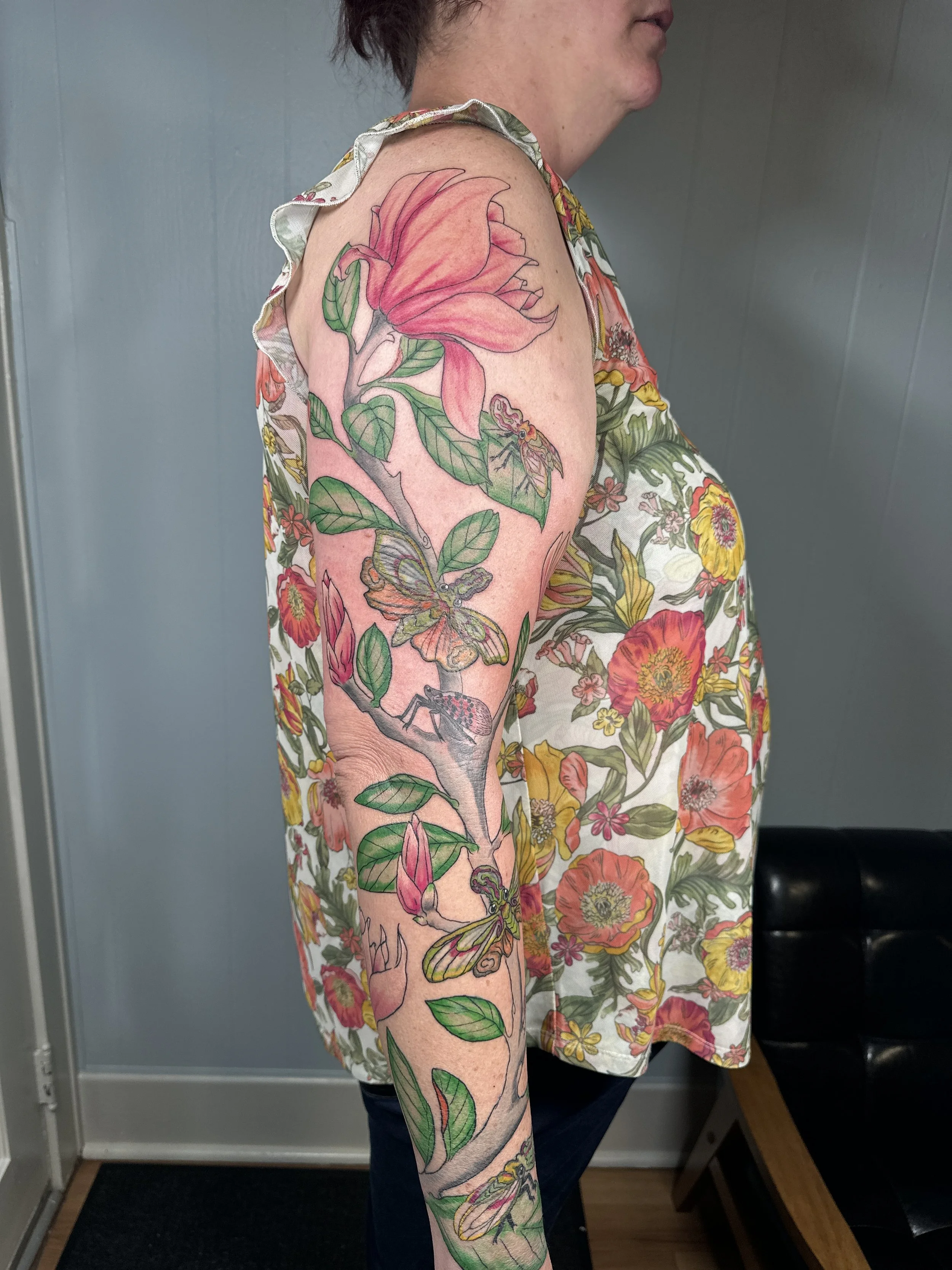 A woman with a floral tattoo sleeve on her upper arm, featuring large pink and peach lily flowers, green leaves, butterflies, a moth, a bee, and other insects, standing in front of a blue wall.