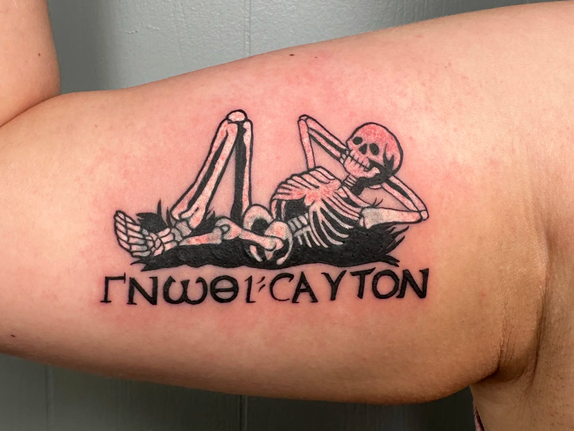 Tattoo of a skeleton lounging on a pillow with the word "ENJOYCATION" underneath it.