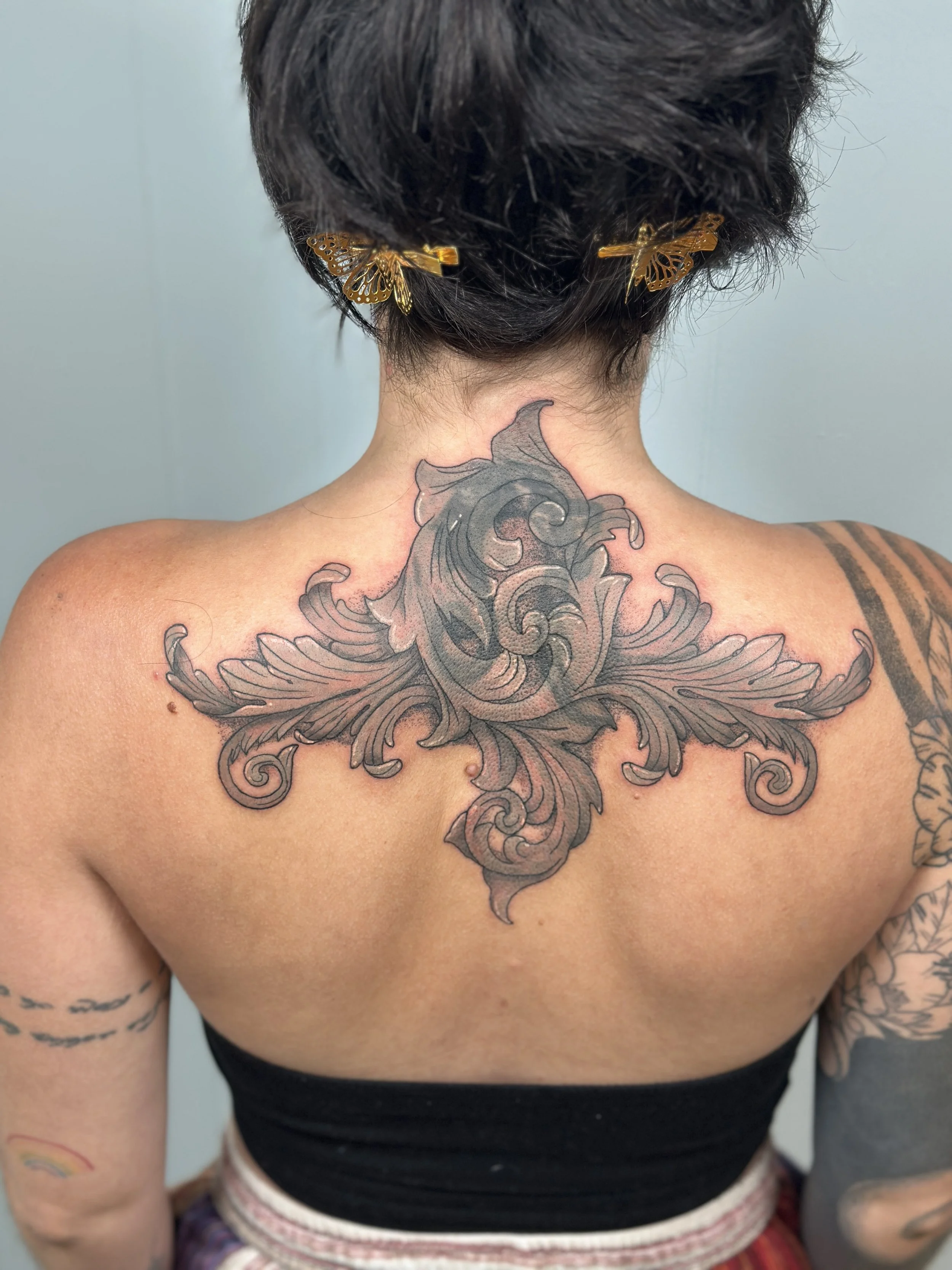 A person with short dark hair wearing butterfly hair clips and a large tattoo on their upper back and shoulders.
