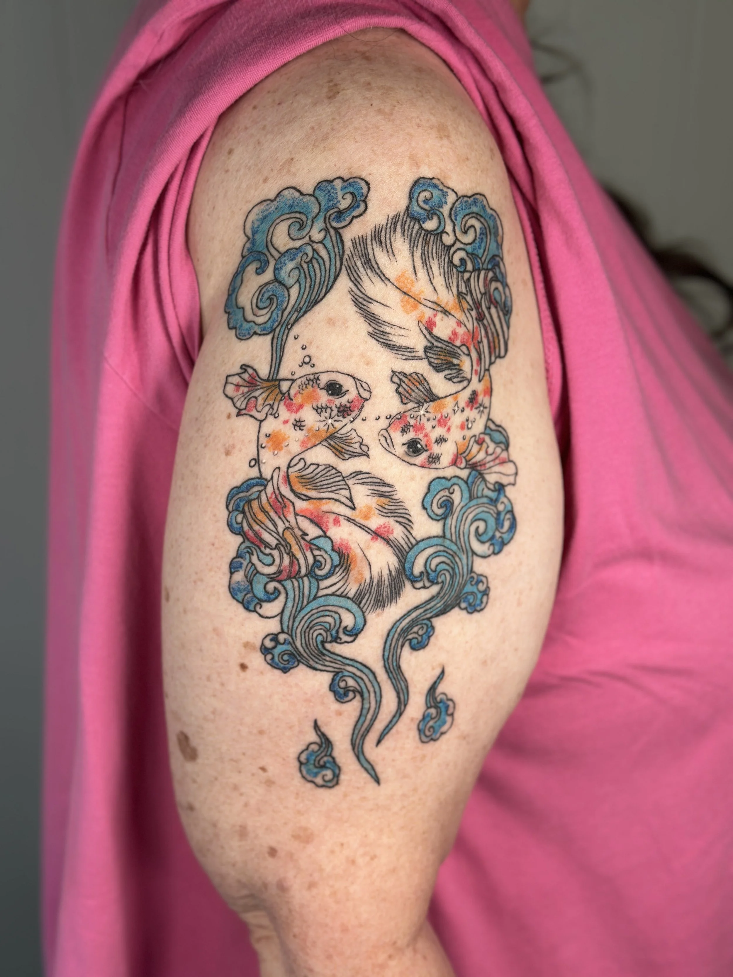 Colorful tattoo of two goldfish surrounded by blue waves on a person's upper arm.