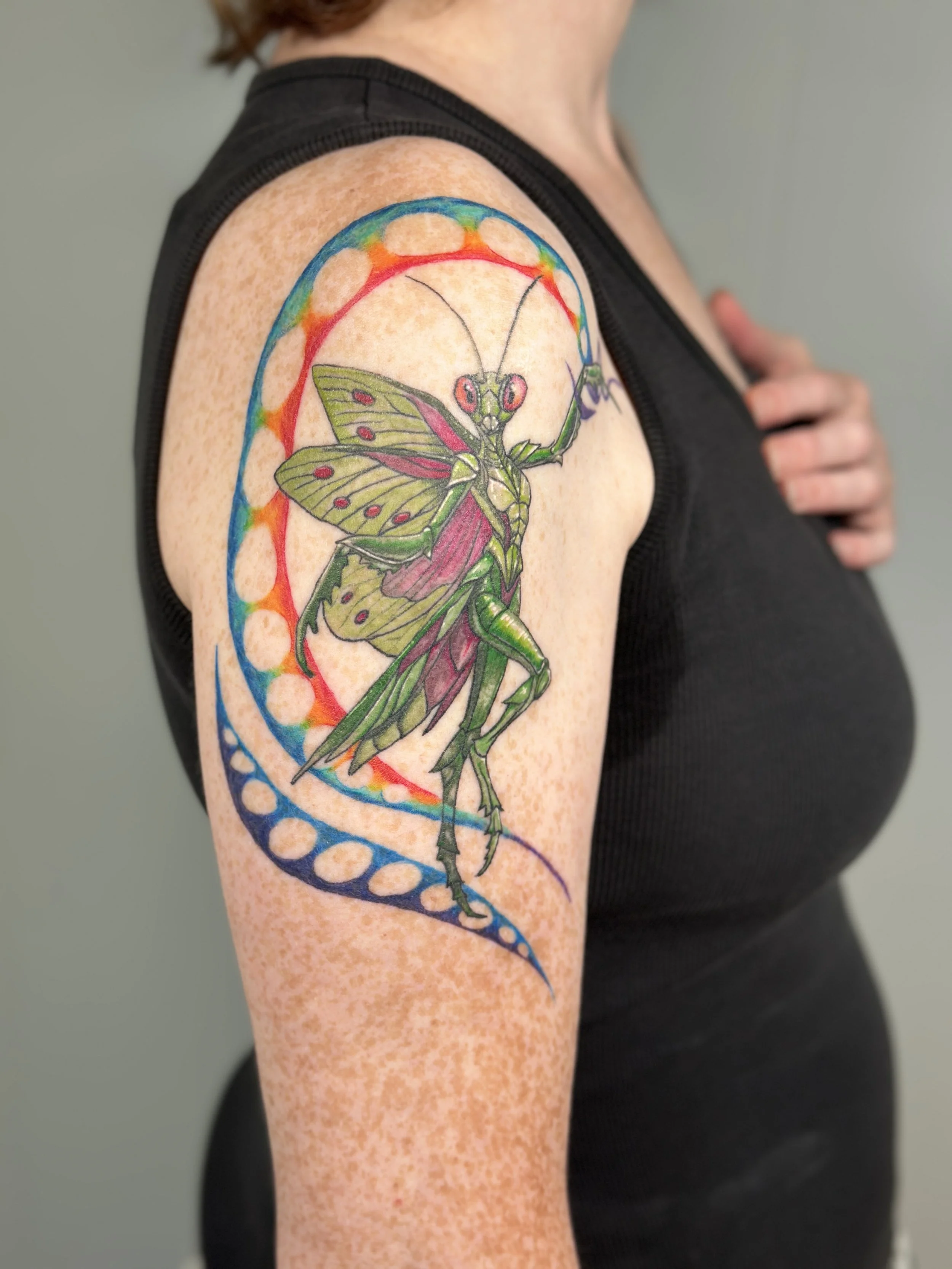 Colorful tattoo of a butterfly with dragonfly wings on upper arm, featuring rainbow-colored circular and curved patterns around it.