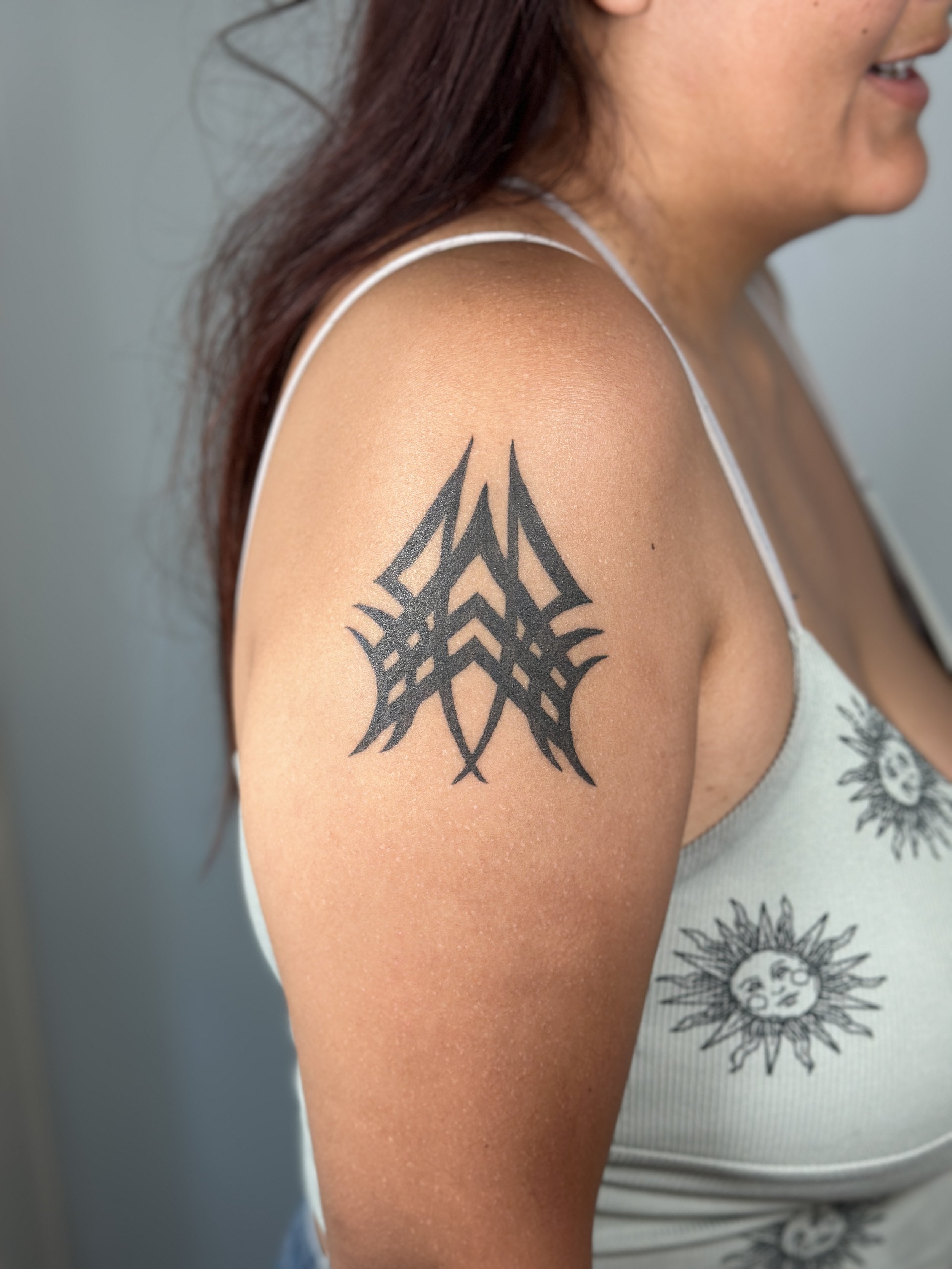 Close-up of a woman's arm and shoulder with a geometric black tattoo, wearing a white tank top with sun and moon designs.