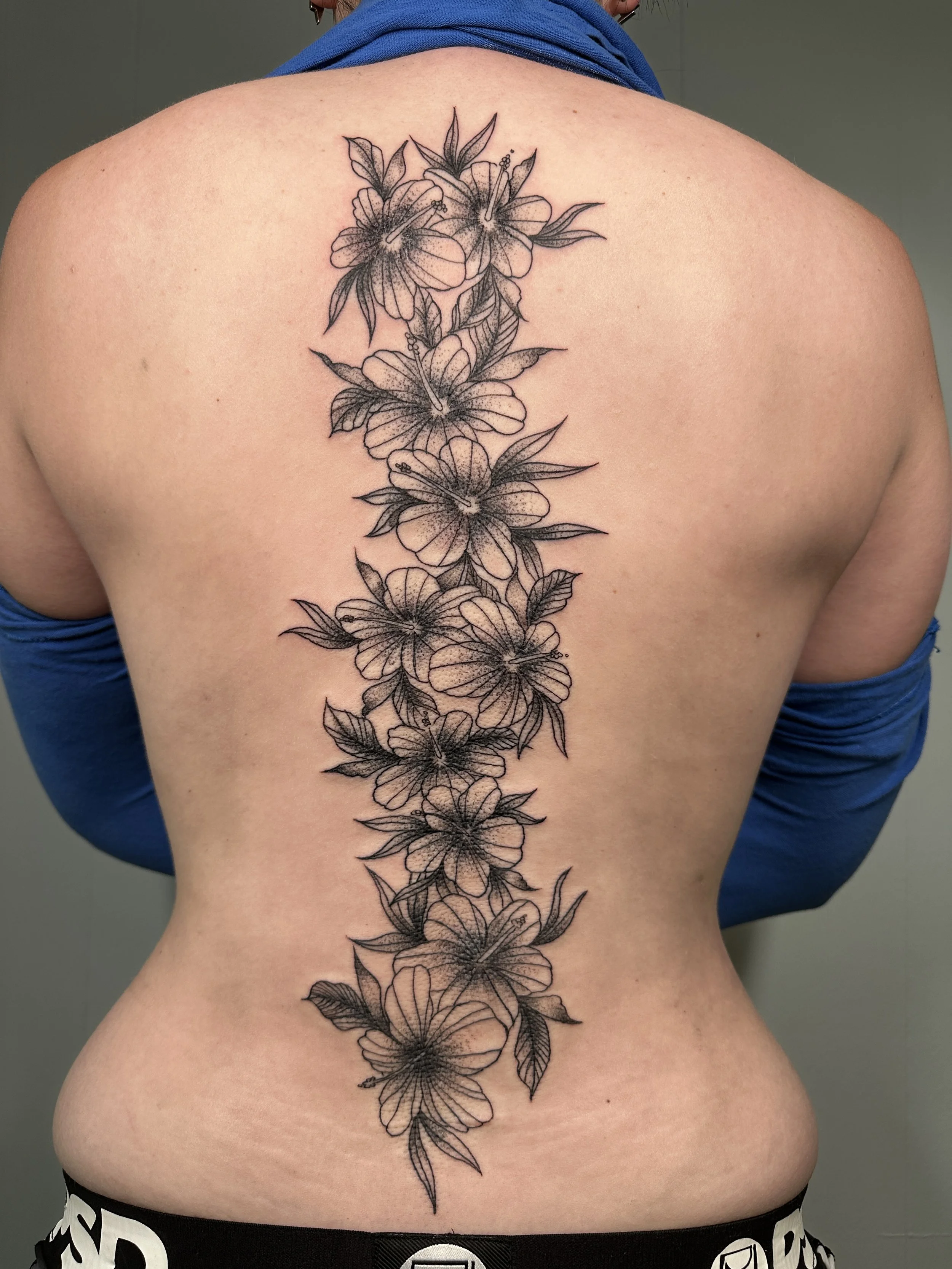 A black and gray floral tattoo of lilies and leaves running down the center of a person's back.