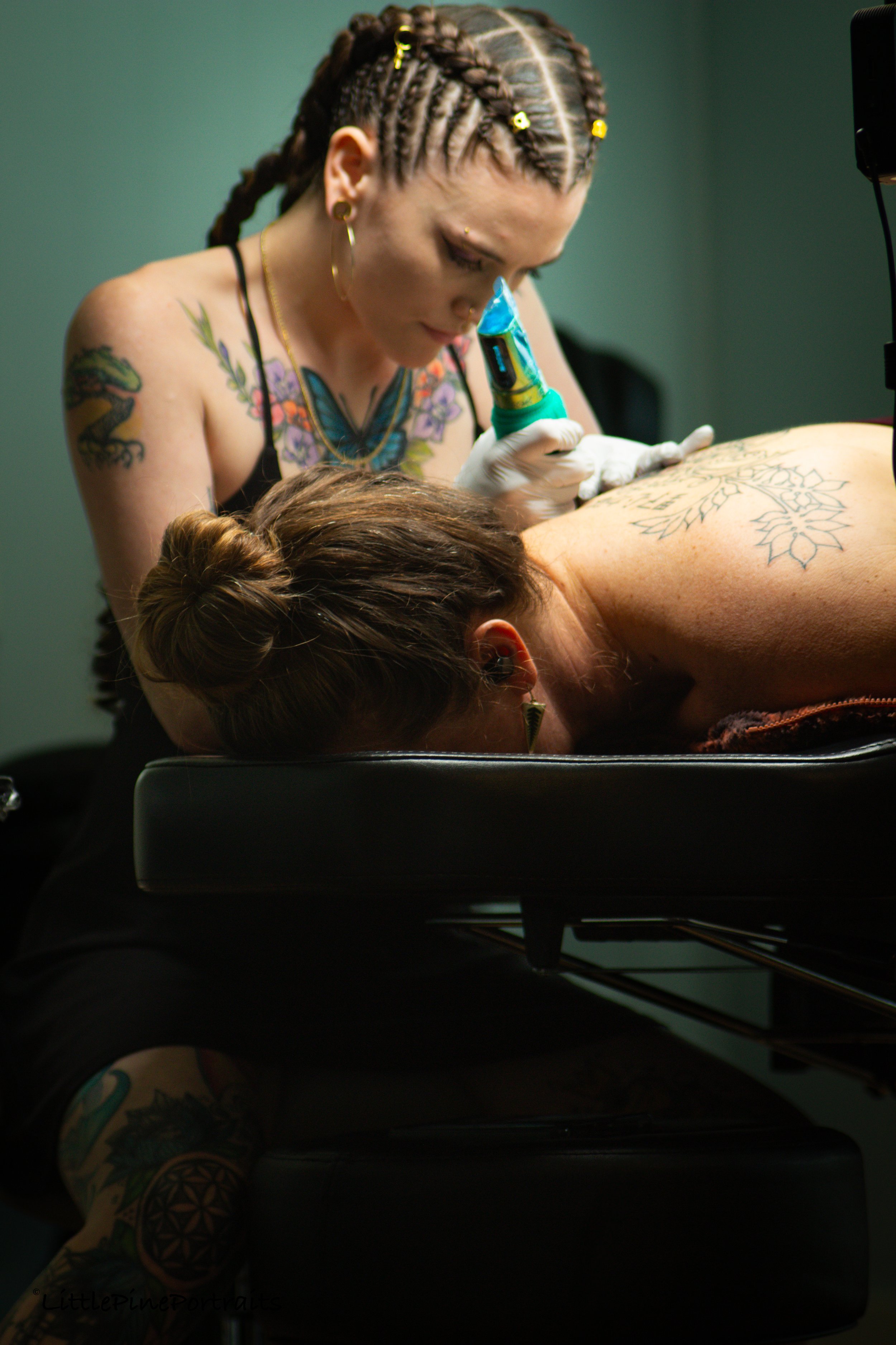 A tattooed woman with braided hair using a tattoo machine on a person's back in a tattoo studio.