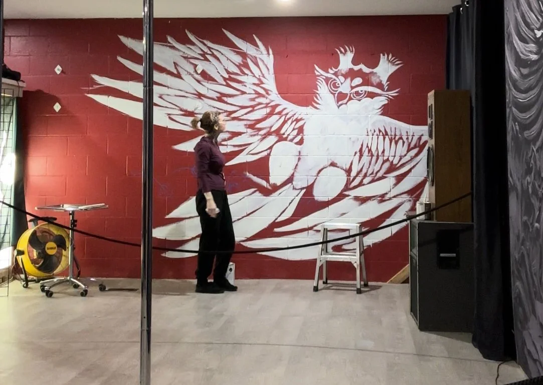A woman standing in front of a large mural of a stylized owl with wings spread wide on a red brick wall, in an indoor setting.
