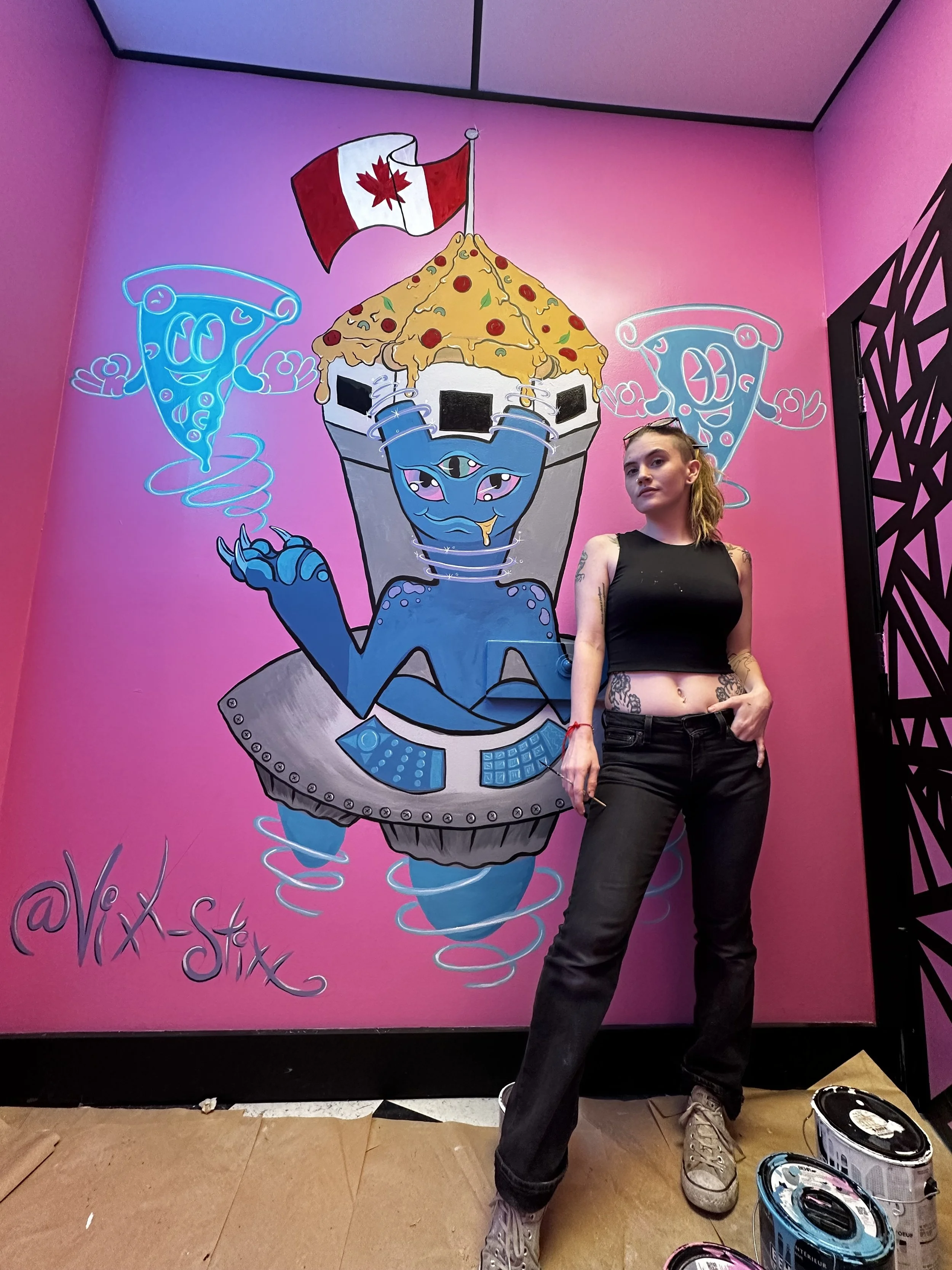 A woman with tattoos dressed in a black sleeveless top and black pants standing in front of a pink wall with a colorful mural of a cartoonish, robotic cupcake with a blue face, multiple eyes, and a cherry topping. The cupcake has a Canadian flag on t