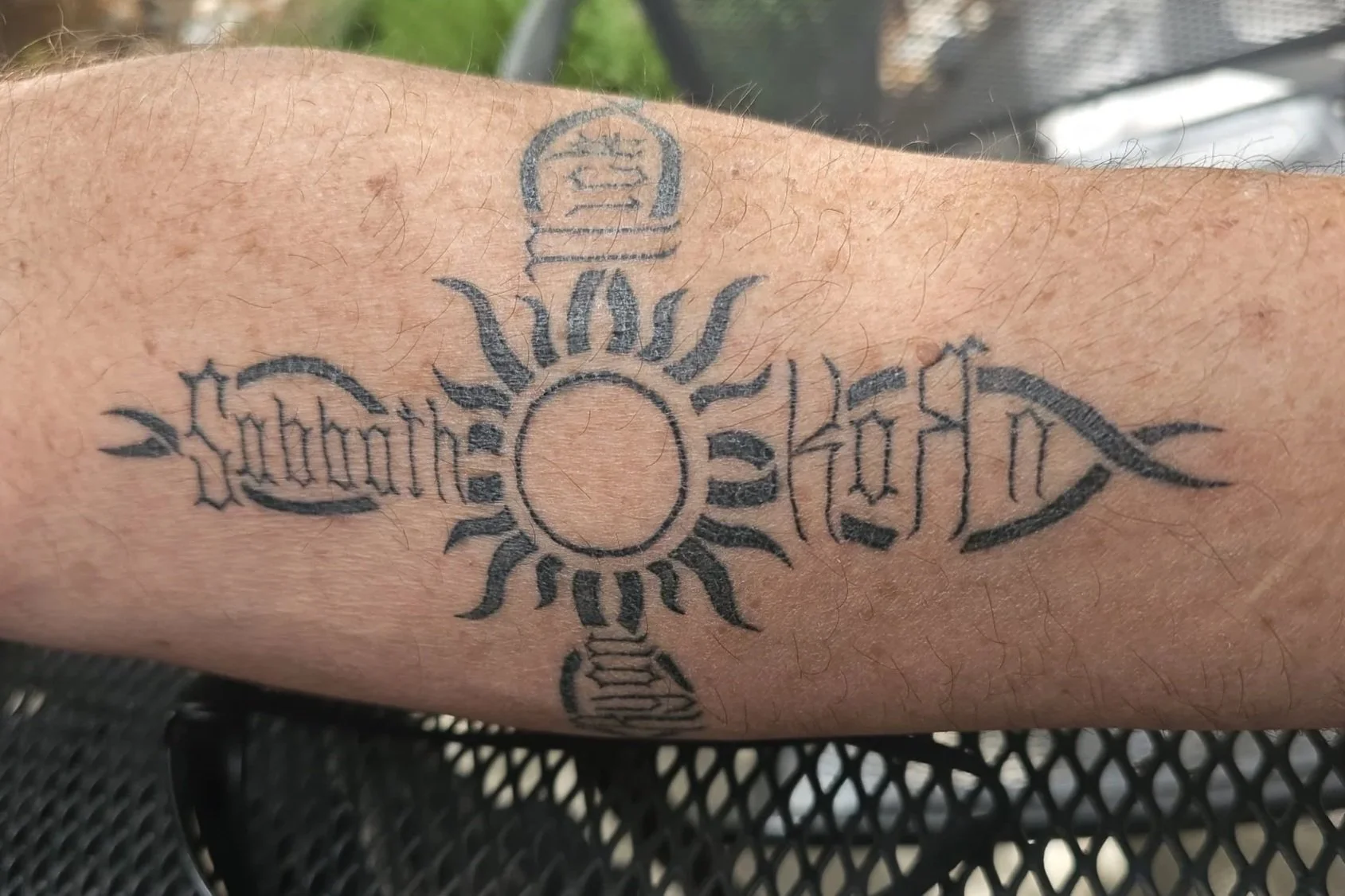 Tattoo of a sun with black rays and a circular center, flanked by the words 'Sabbath' on the left and 'Holy' on the right, on a person's arm.