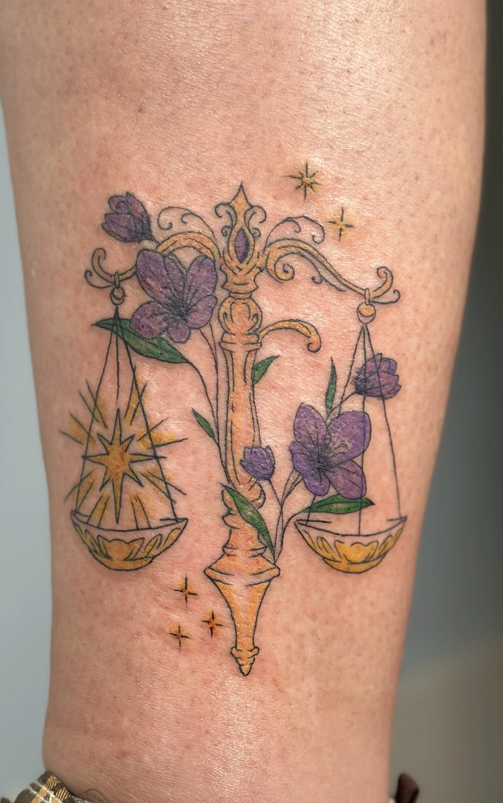 Colorful tattoo of a balanced scale with purple butterflies, a decorative central column, and small stars on light skin.