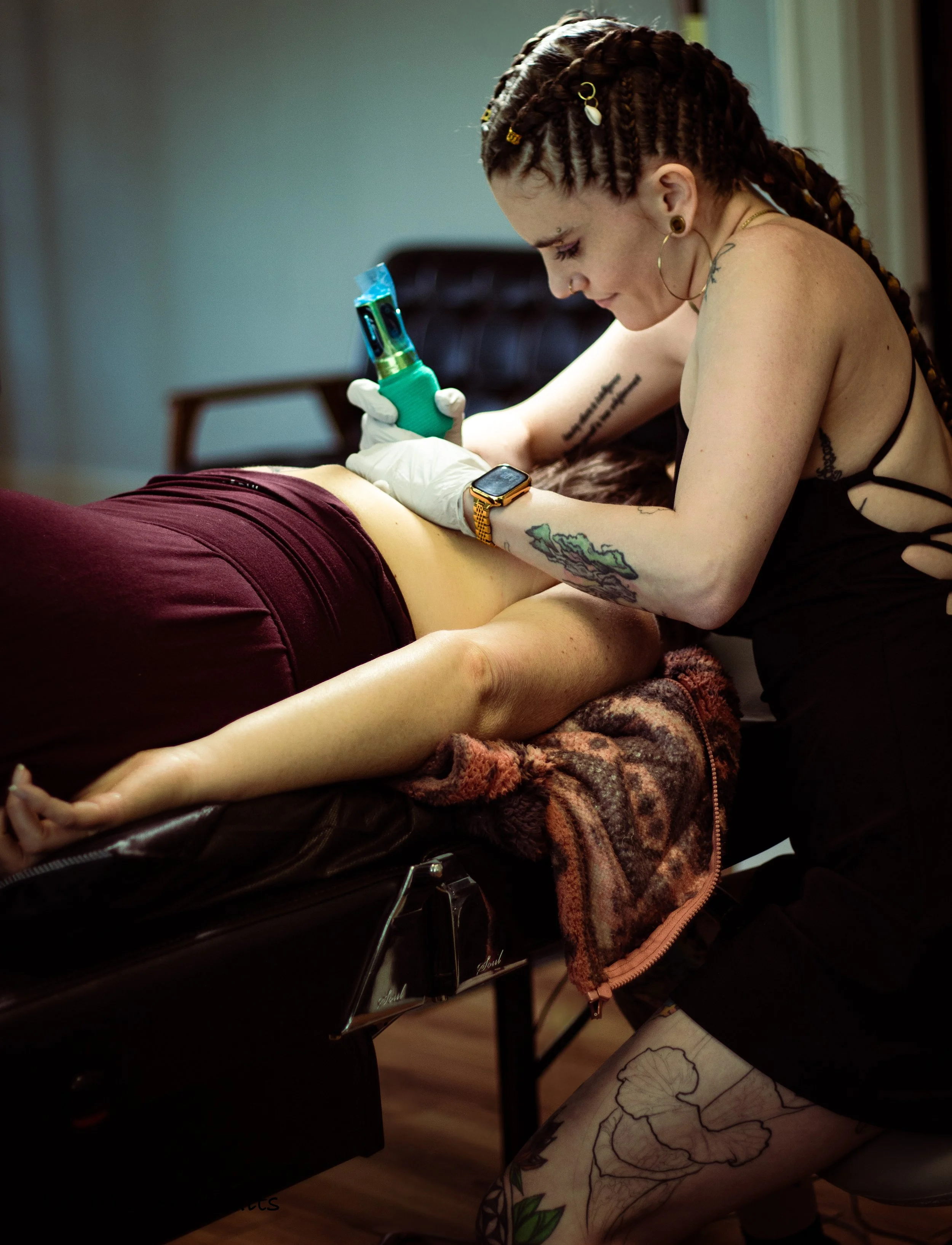 A tattoo artist with braided hair, multiple earrings, and tattoos on her arm, wearing earrings and a watch, is tattooing a floral design on a person's leg. The person is lying on a black tattoo bed covered with a blanket, and their leg is exposed.