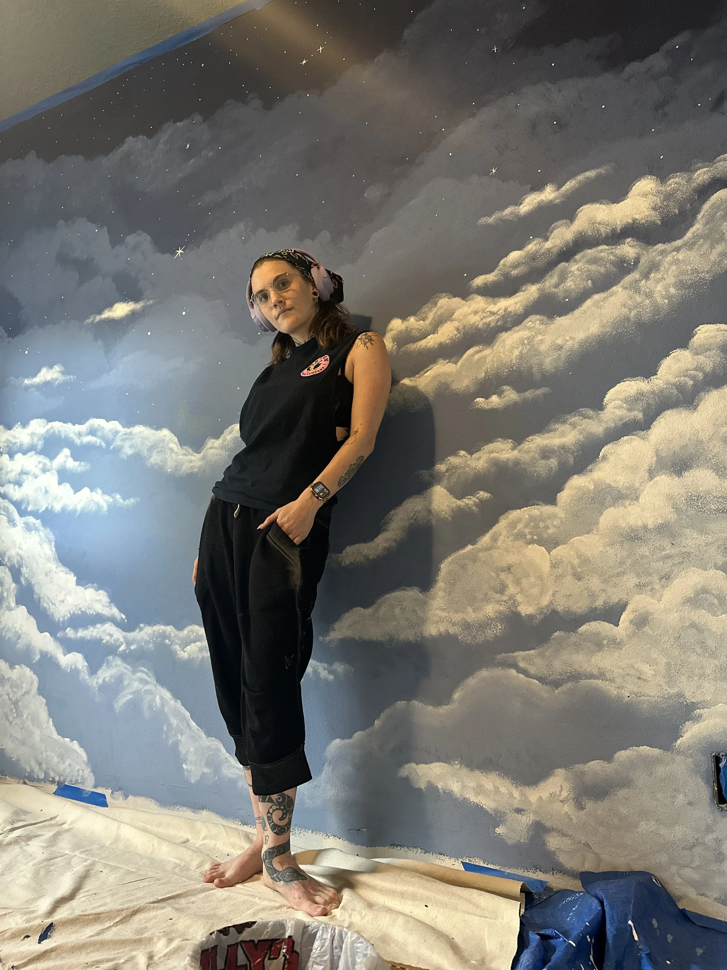 Woman with tattoos, wearing glasses, a headscarf, and headphones, standing on a beige sheet in front of a backdrop painted with clouds and stars, resembling the sky.