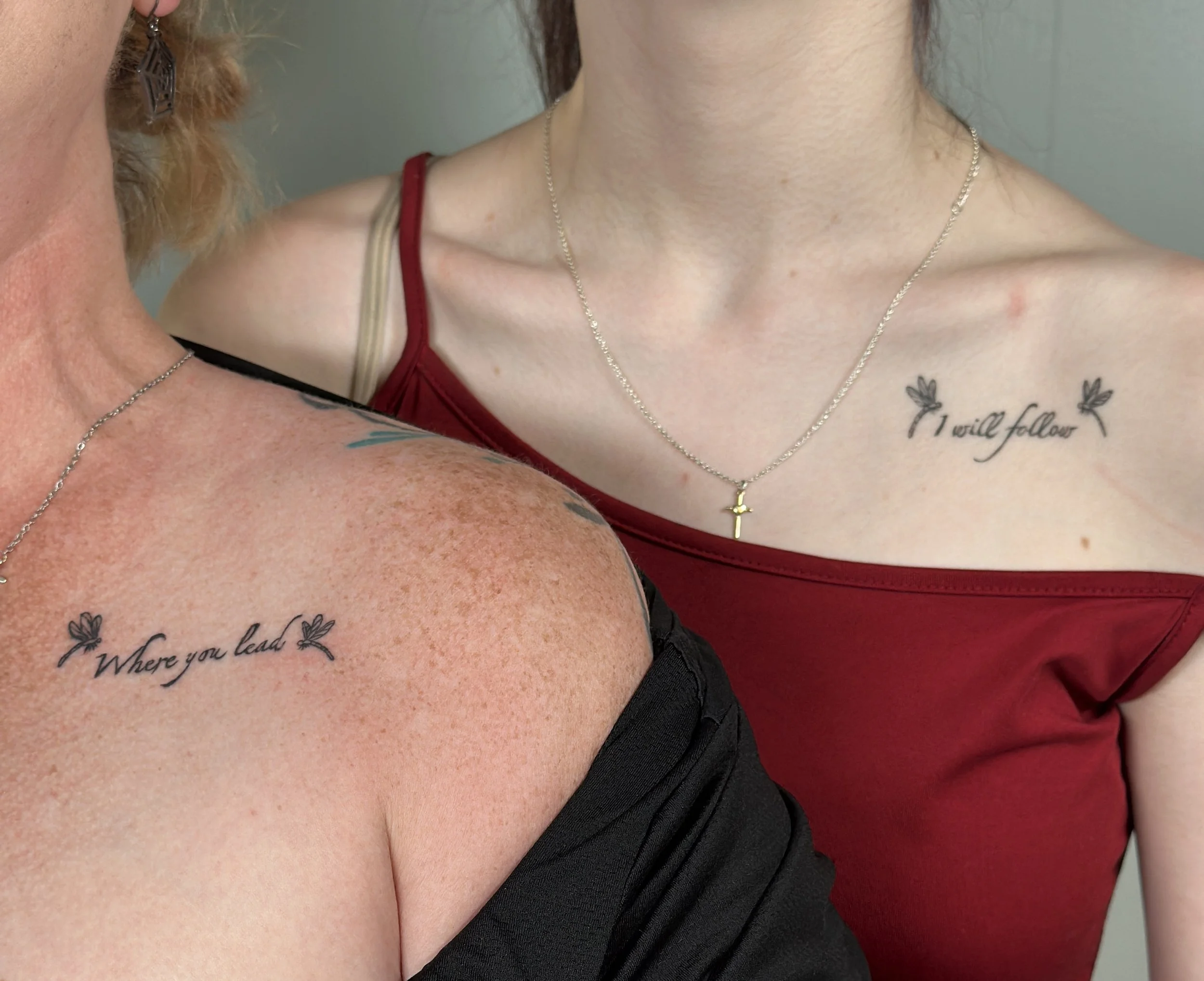 Close-up of two women showing their shoulders and necklaces, with visible tattoos and clothing, in a casual setting.