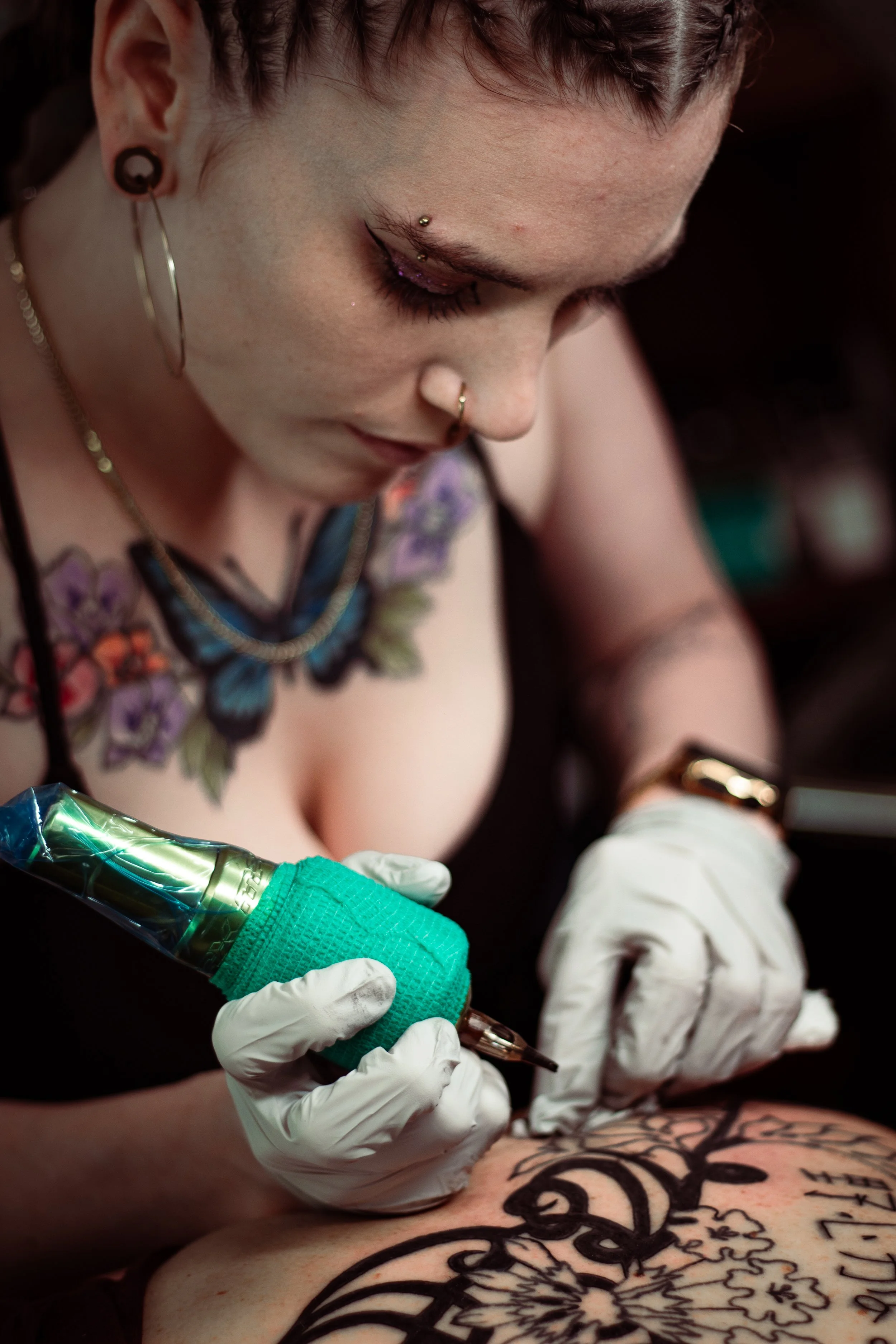 A person with tattoos reciving a tattoo from an artist wearing-gloves, holding a tattoo machine, on their back.