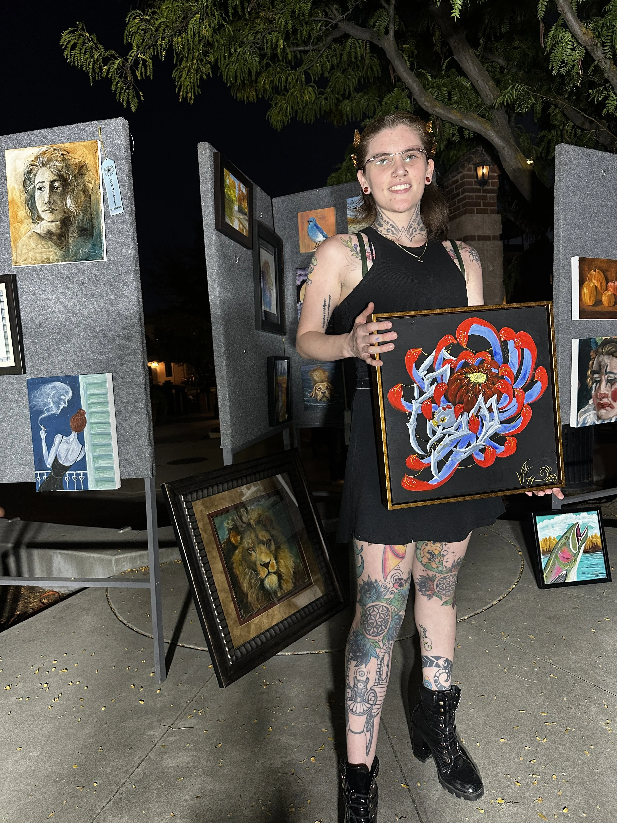 A young woman with numerous tattoos, glasses, and black boots is holding a colorful painting of a flower at an outdoor art display at night. She is smiling and standing amidst various framed artworks, including portraits of a lion, a woman, and other