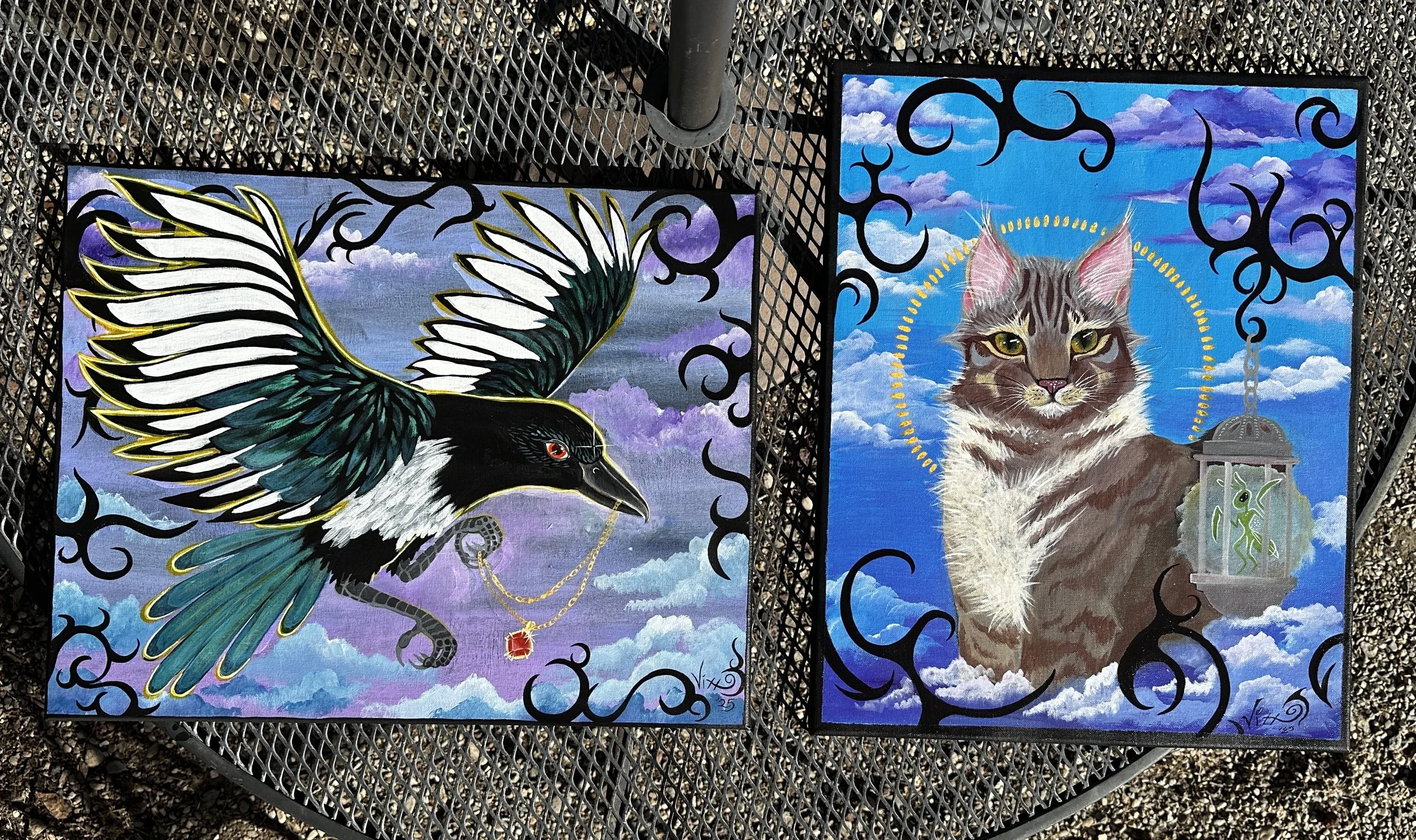 Two colorful paintings on a black metal mesh table. The left painting depicts a bird with black and white feathers, a yellow outline, a blue background, clouds, and a red jewel hanging from a chain. The right painting shows a cat with green eyes, bro