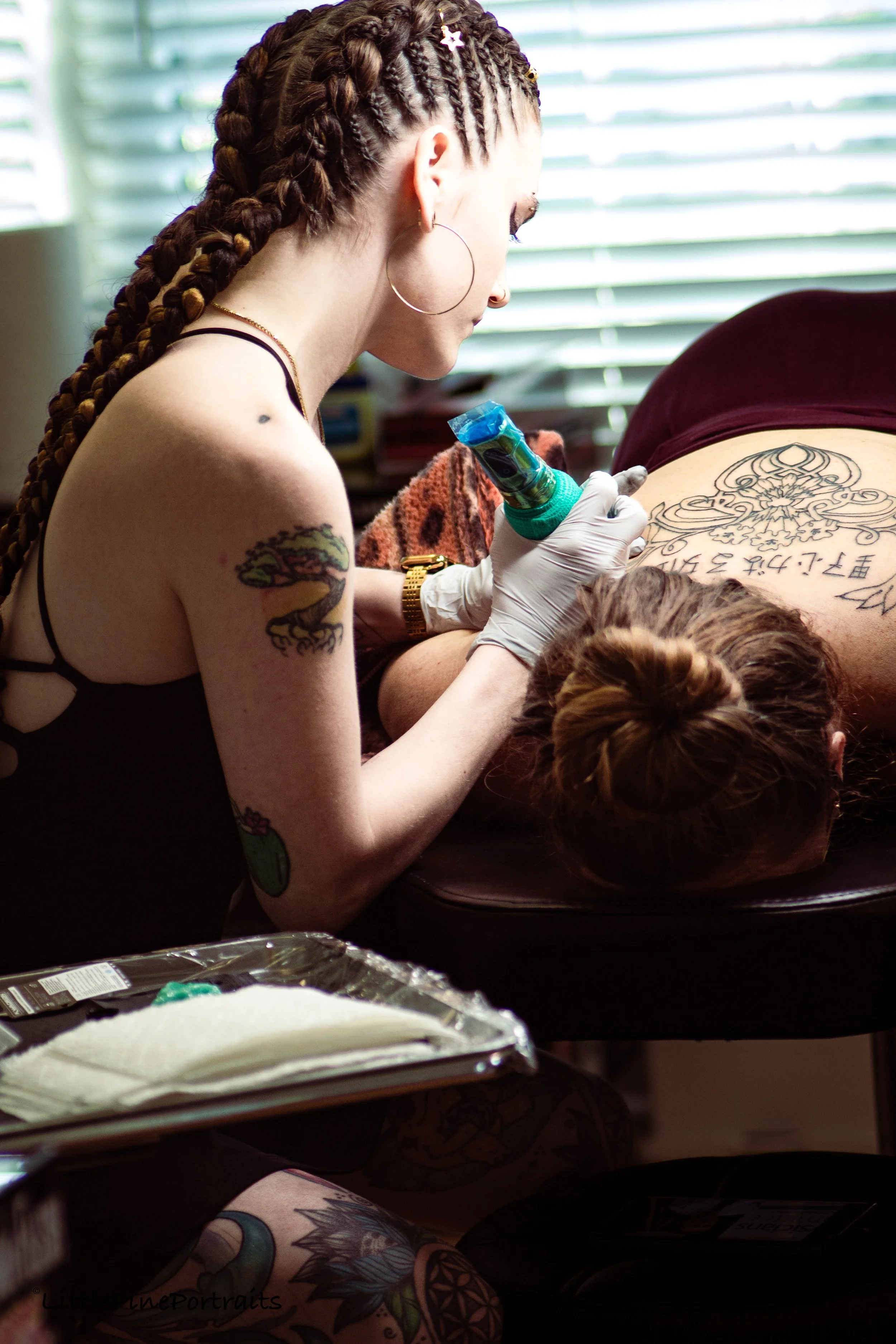 A tattooed woman with braids is tattooing a large, ornate design with Chinese characters on a person's back. She is wearing a black top, hoop earrings, and a gold watch, and is holding a tattoo machine.