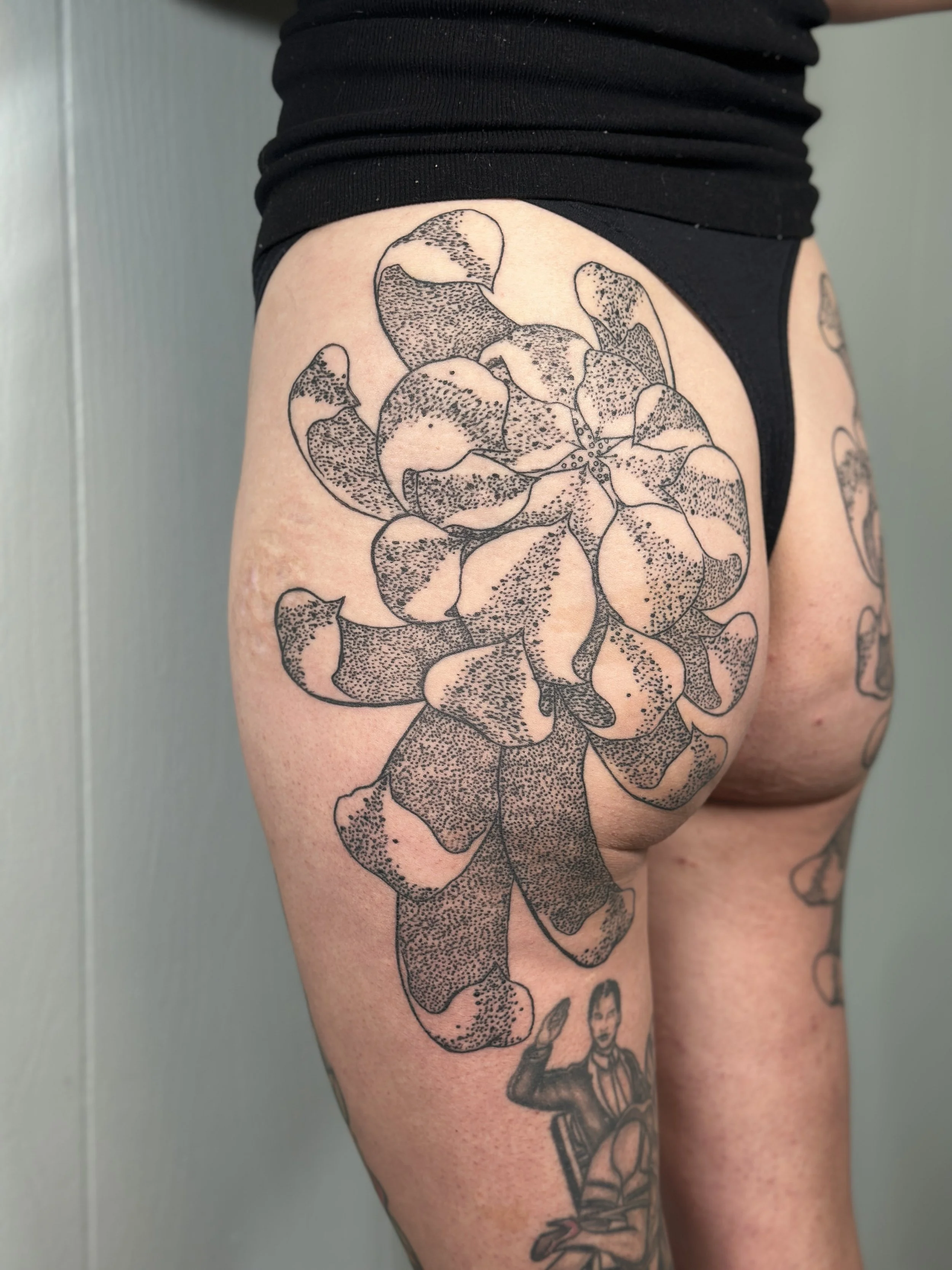 Black line-drawing tattoo of a large flower, possibly a rose, on a person's thigh, with additional tattoos visible on the leg and other parts of the body.