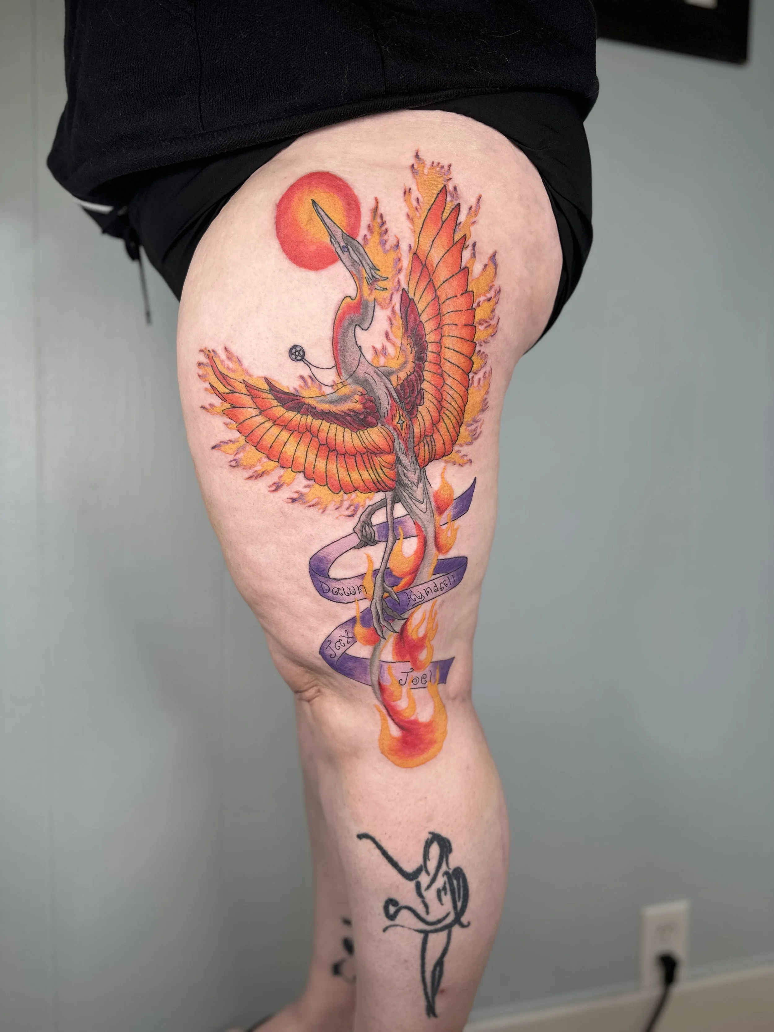 Tattoo of a phoenix with flames, a sun, and text ribbons on a person's thigh.