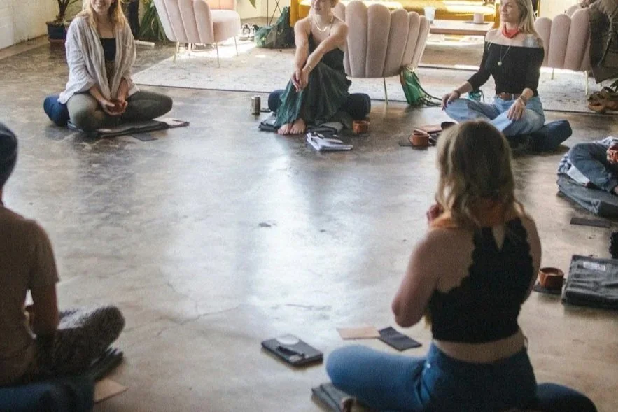 A group of young women sitting on the floor in a circle during a gathering or workshop in a bright, airy room with modern decor.