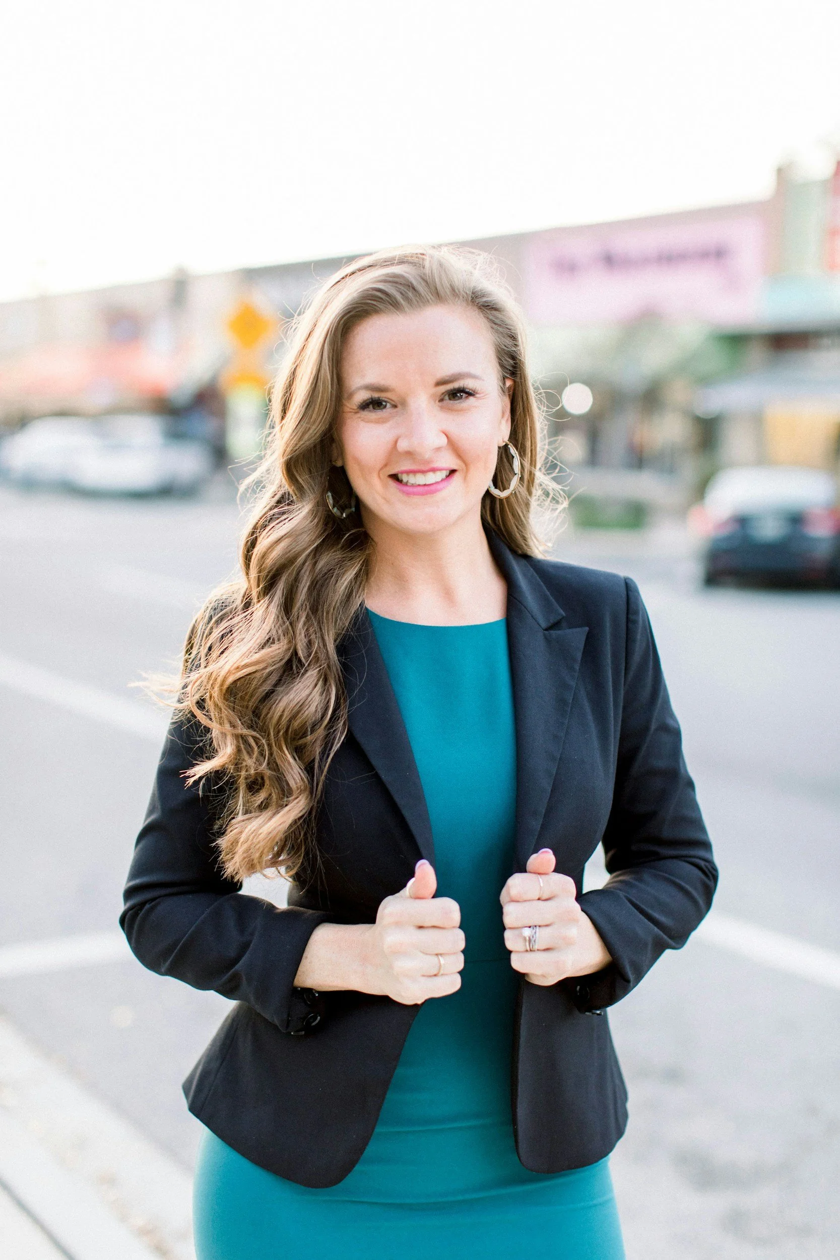 A woman with long wavy hair wearing a dark blazer and teal dress, smiling and standing outdoors in a parking lot.