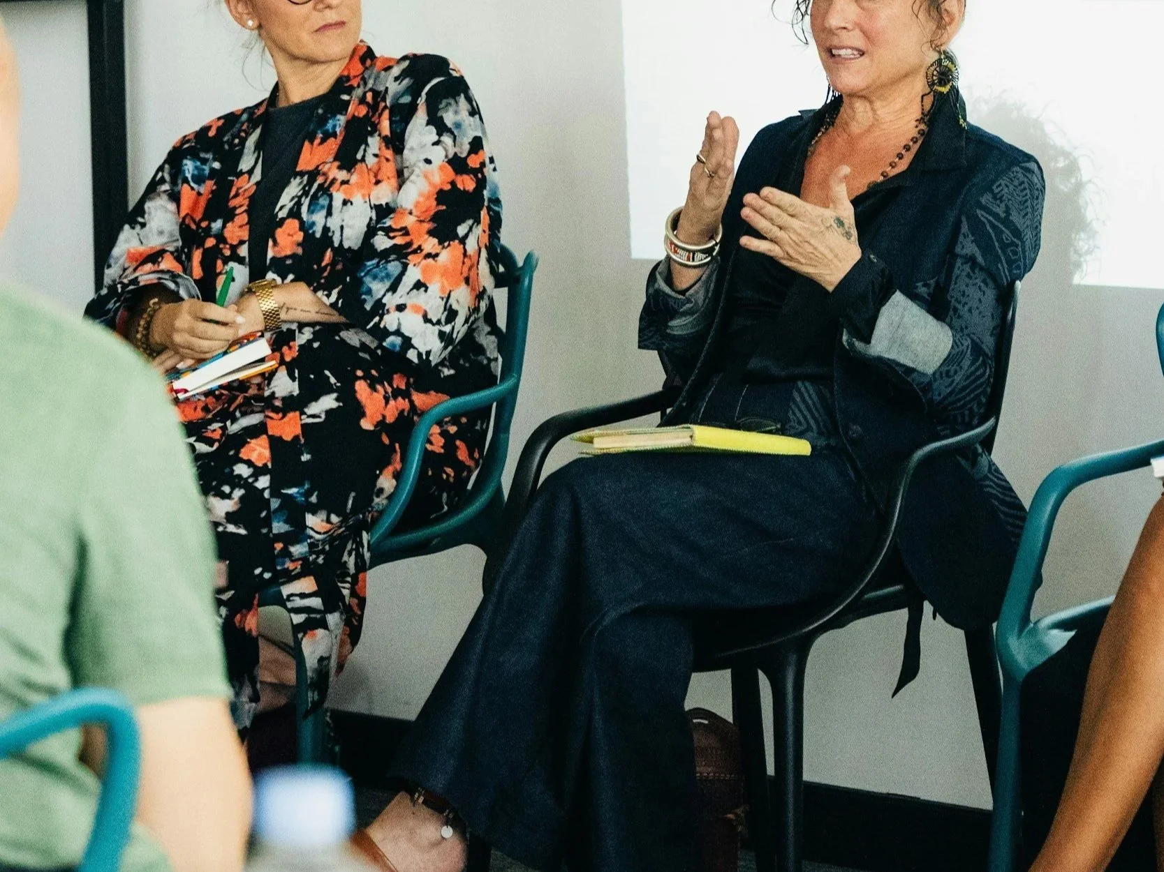 Two women sitting in a meeting, engaged in conversation. The woman on the right is speaking and gesturing with her hand, while the woman on the left is listening and holding a notepad. Both are wearing dark and patterned clothing, with one having earrings and bracelets.