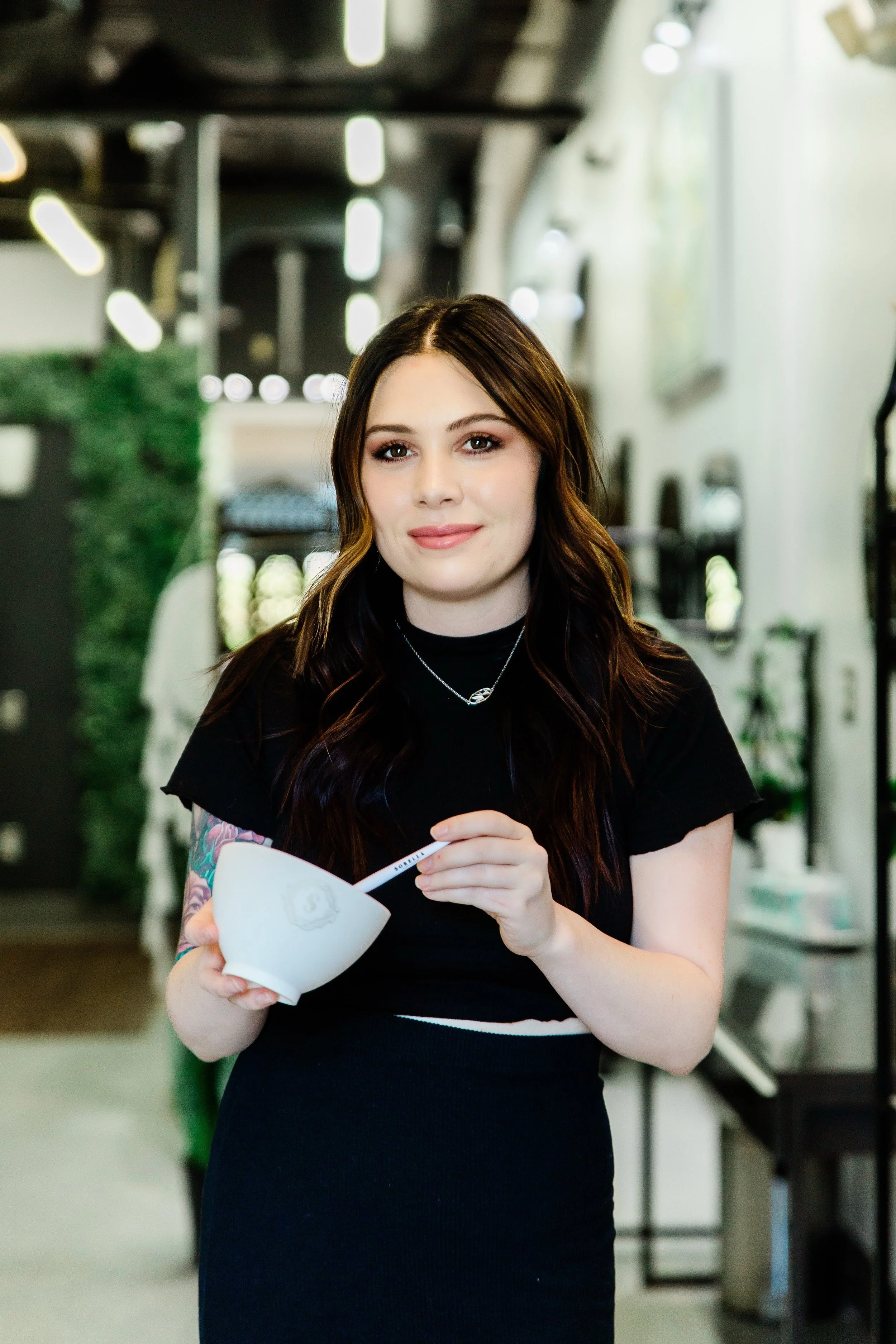 A young woman with long brown hair and light makeup, wearing a black dress and a silver necklace, holds a white bowl and a spoon, standing in a modern indoor setting with blurred background elements.