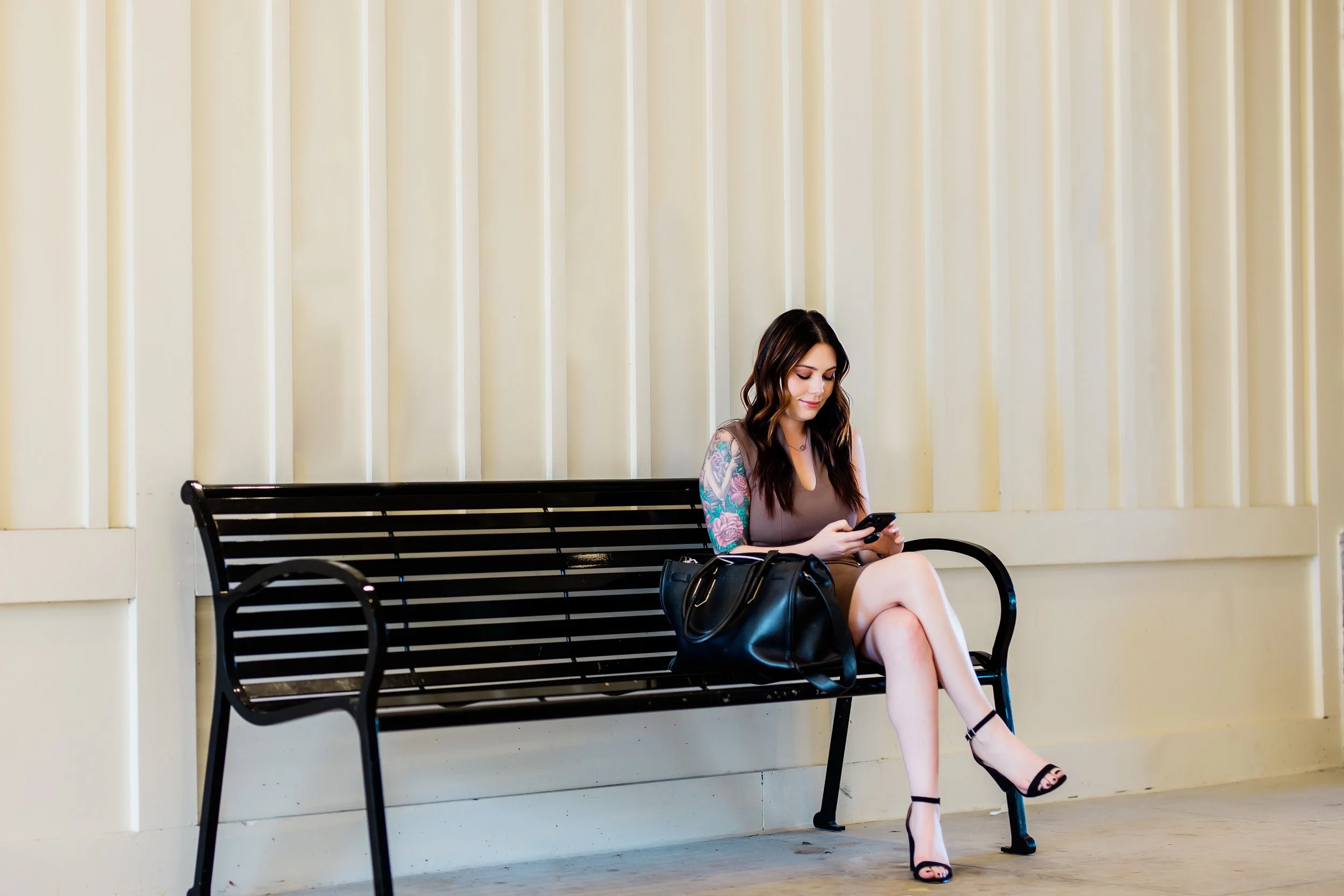 A woman with long dark hair and tattoos on her arm sitting on a black metal bench, looking at her phone, wearing a beige dress and black high-heeled sandals, with a black purse beside her, in front of a beige wall with vertical panels.