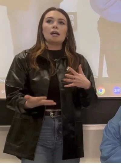 A woman with medium-length brown hair wearing a black jacket over a black top, standing in front of a whiteboard in a classroom or conference room, speaking and gesturing with her left hand on her chest.