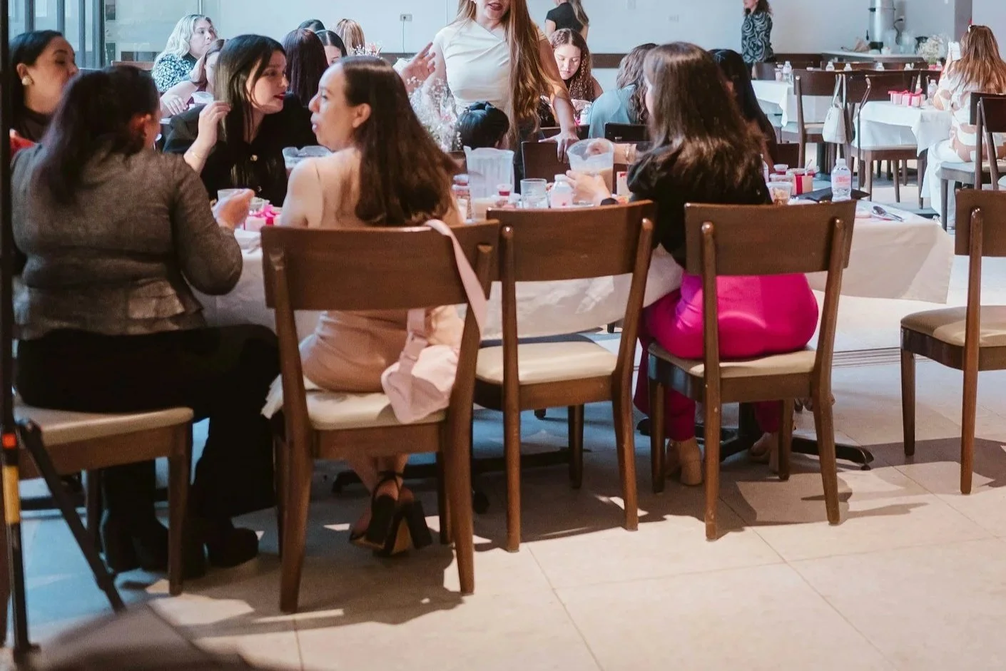A group of women sitting around a table at a social gathering or celebration, engaged in conversation, in a well-lit indoor setting with other tables and women in the background.