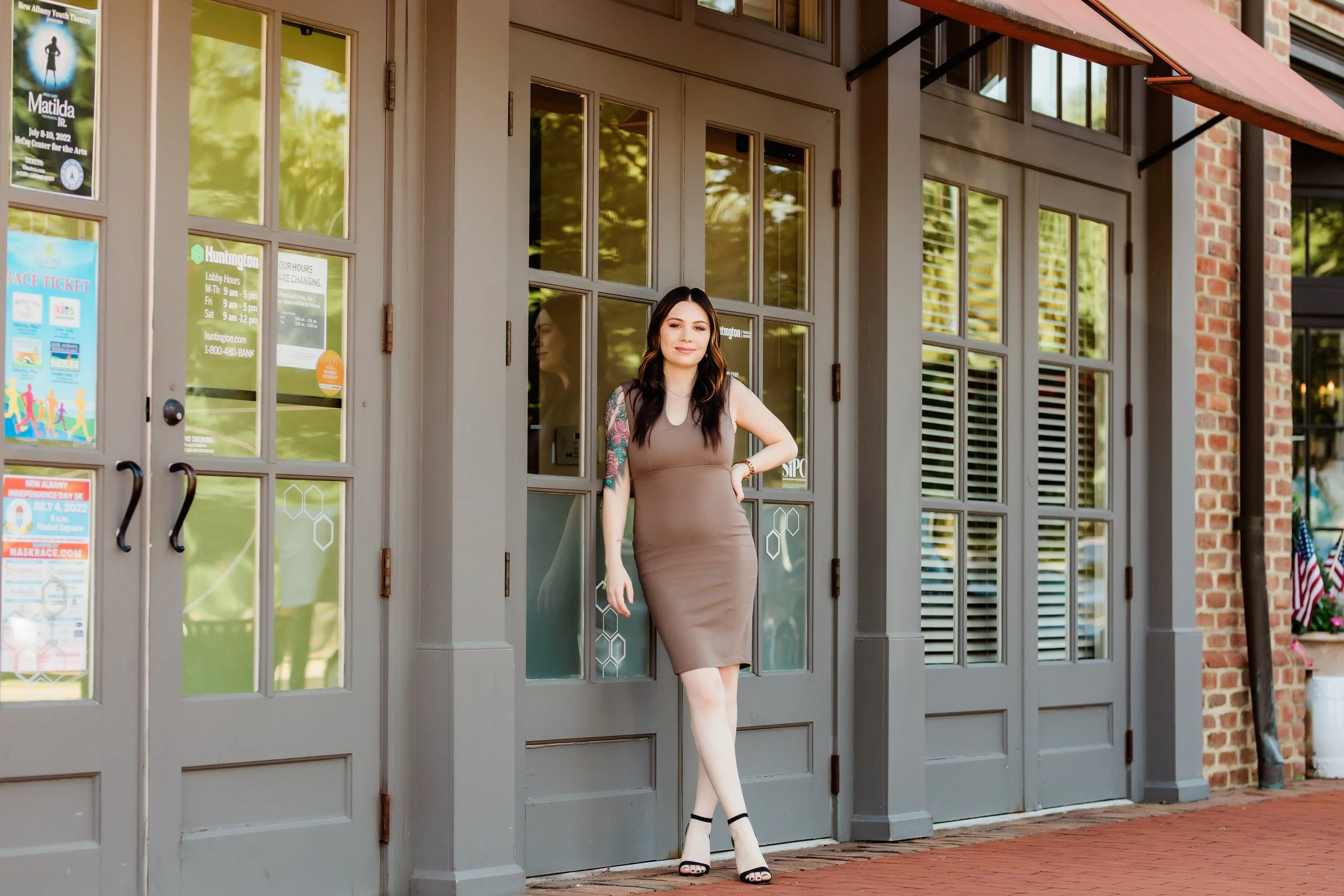 A woman standing at the doorway of a building with large glass-paned doors. She is wearing a form-fitting, sleeveless, taupe-colored dress and black high-heeled sandals. She has long dark hair, a tattoo on her right arm, and is smiling at the camera. The building has a brick wall to the right and a small American flag and potted flowers near the entrance.