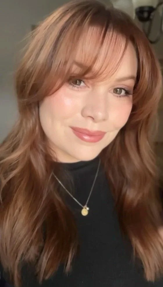 Close-up of a woman with long, wavy, reddish-brown hair, wearing a black top and a gold pendant necklace, smiling at the camera.