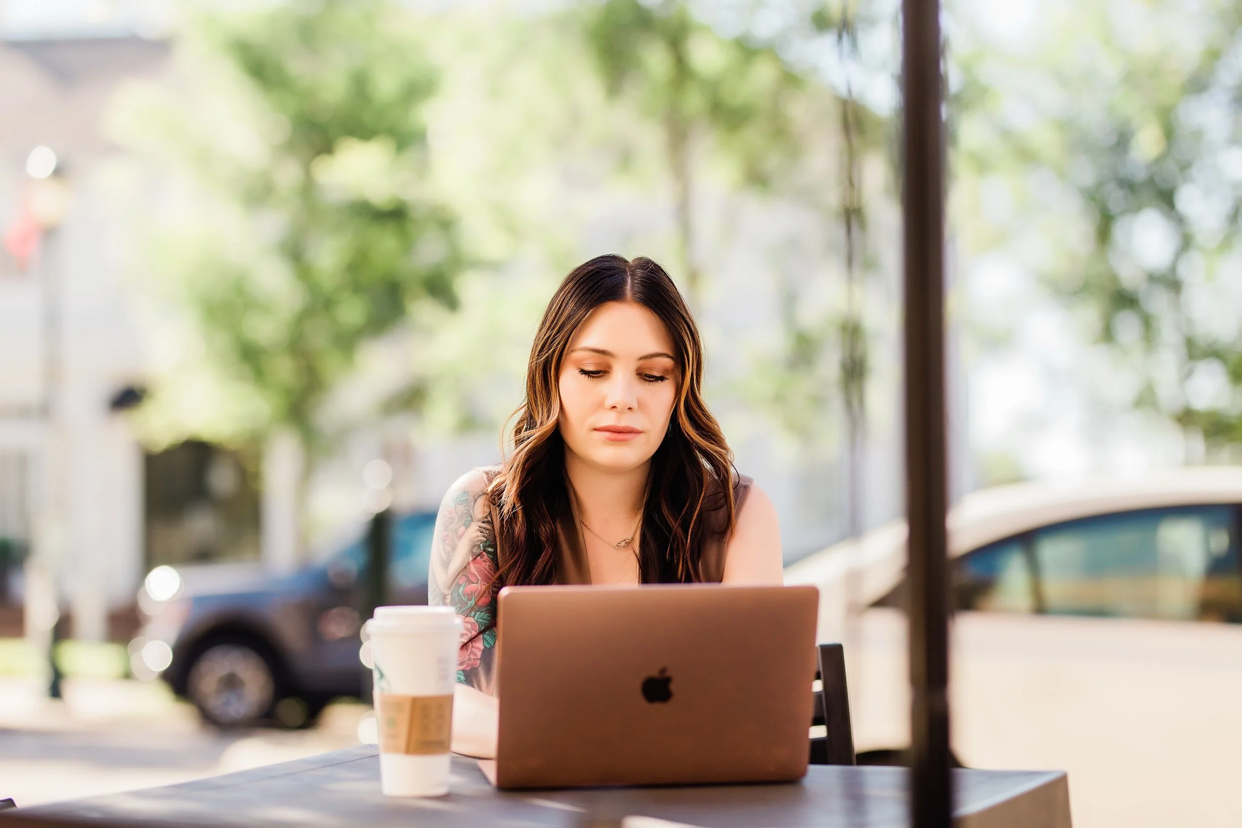 A young woman with long, wavy brown hair and tattoos on her arms sitting at an outdoor table, looking at her laptop. Coffee cup and smartphone on the table. Green trees and parked cars in the background.