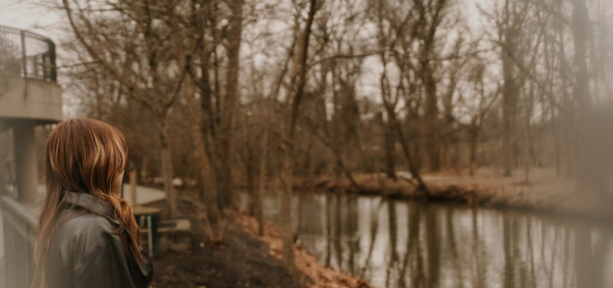 A woman with long brown hair wearing a gray jacket looking out over a river in a wooded area during autumn.