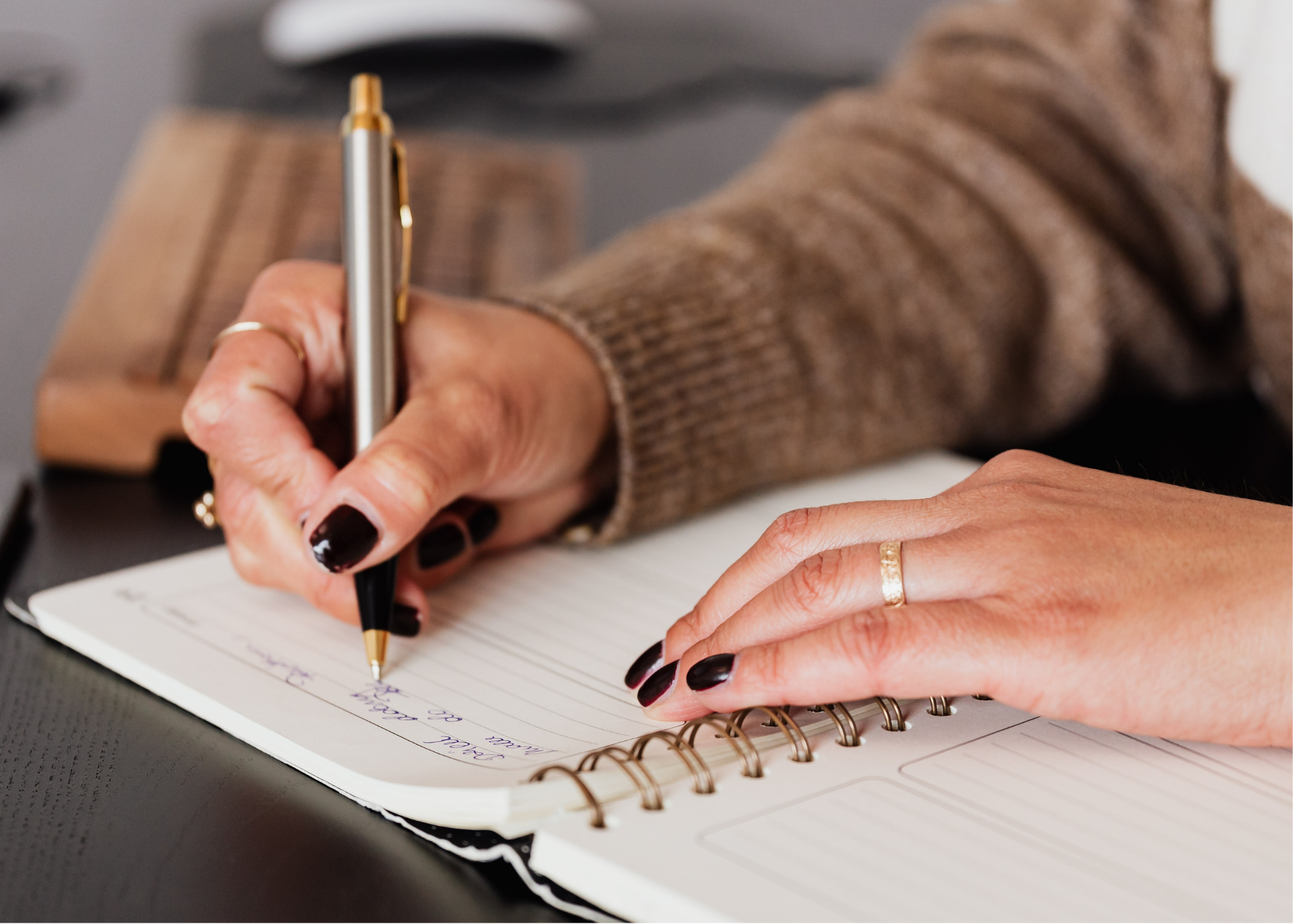 Close-up of a person's hand writing in a spiral notebook with a black and gold pen. The person is wearing a brown sweater and a gold ring. The notebook contains handwritten notes.