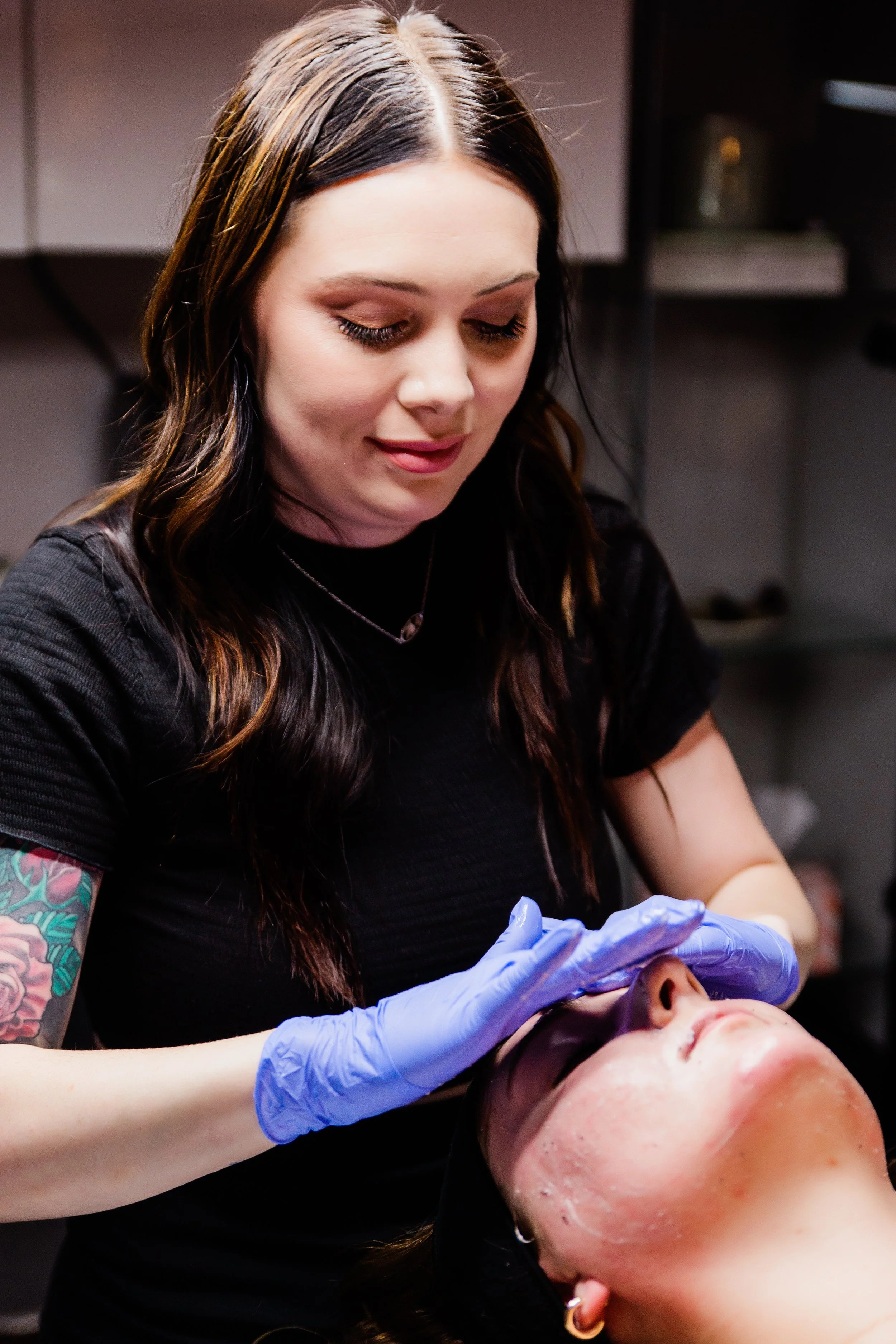 A tattooed woman with long wavy hair, wearing black, is performing a facial treatment on a woman lying down with closed eyes. She is wearing blue gloves and is in a professional setting.