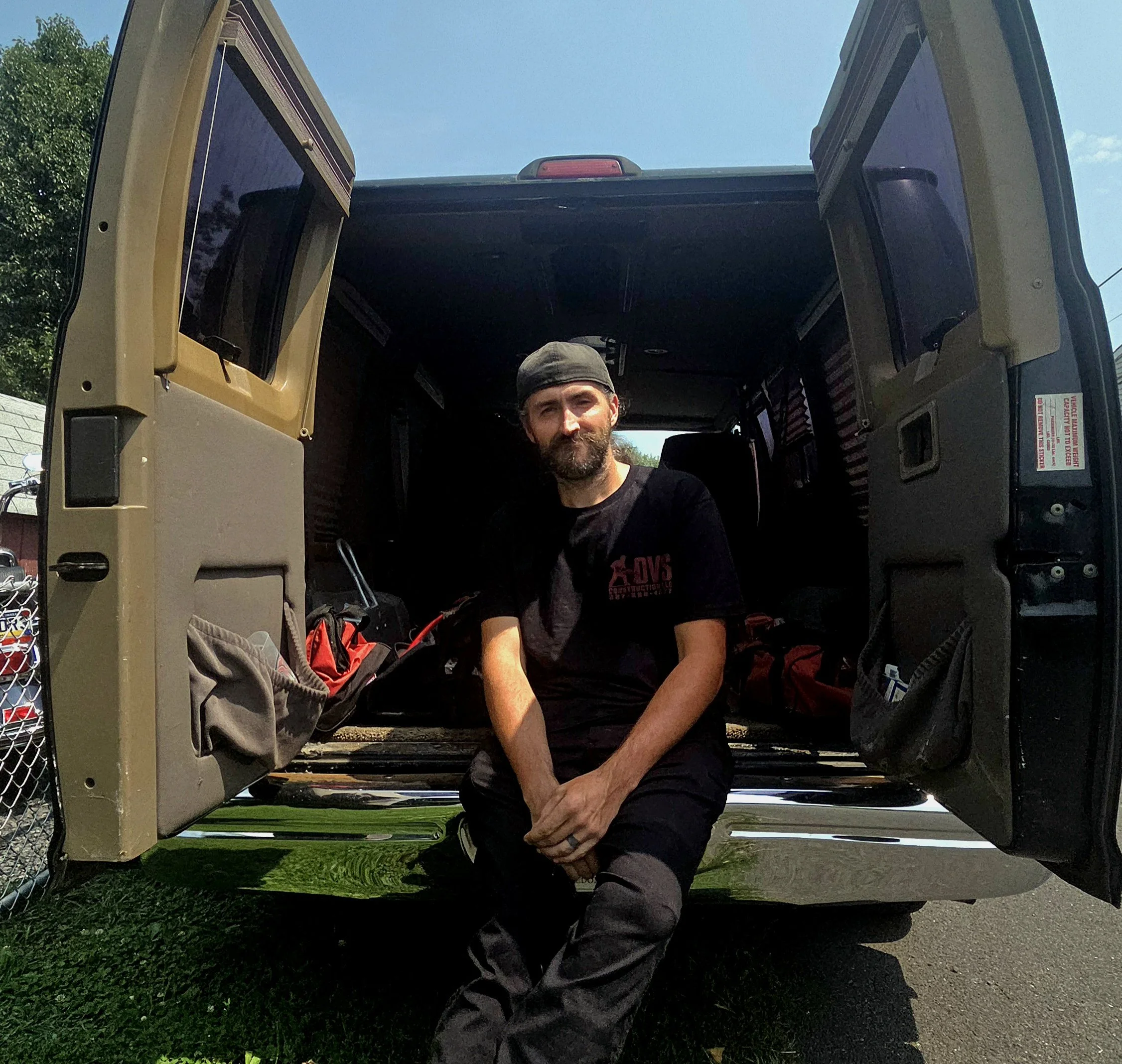 A man with a beard and black cap sitting inside an open door of a utility or military vehicle, smiling at the camera, with a blue sky and green trees in the background.