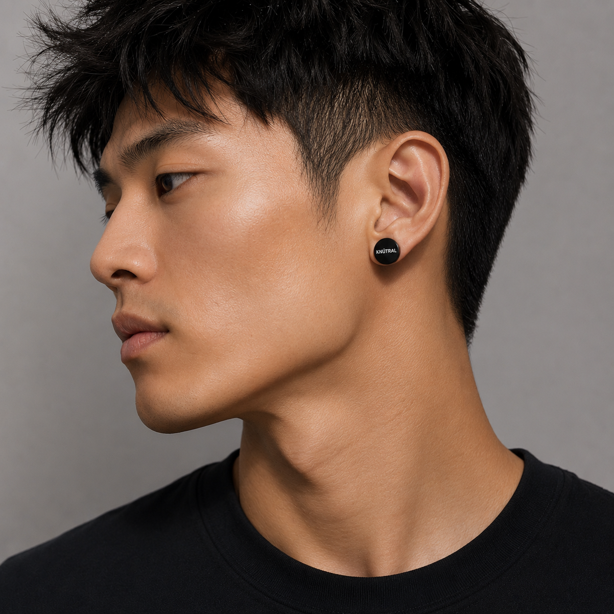 Close-up side profile of a young man with short black hair, wearing black earbuds labeled 'KNUTRAL', against a gray background.
